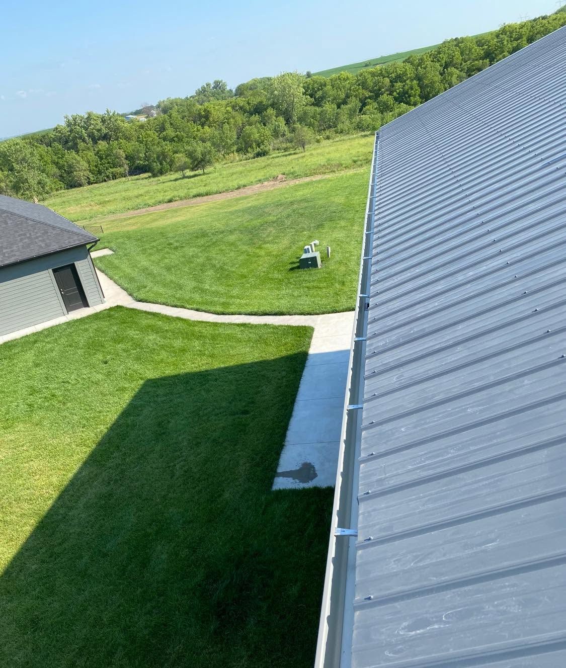 View from a rooftop over green lawn, walkway, and distant trees beside a metal roof.