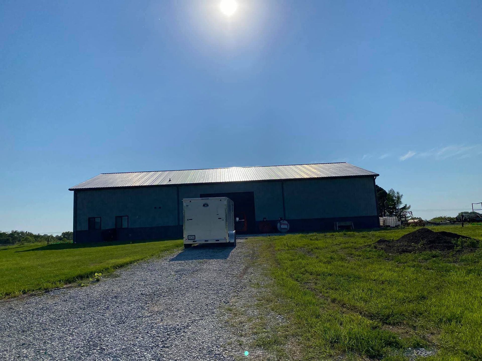 A long dark barn with a white roof sits beside a gravel drive under a bright blue sky.