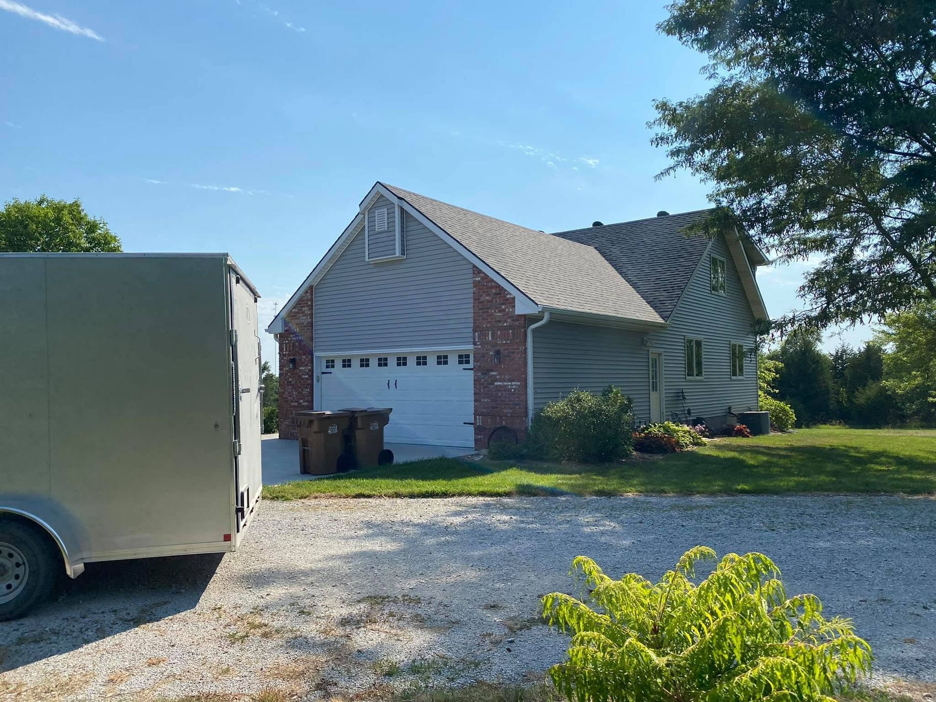Suburban house with white garage, gravel driveway, and a trailer parked on the left under a clear blue sky.