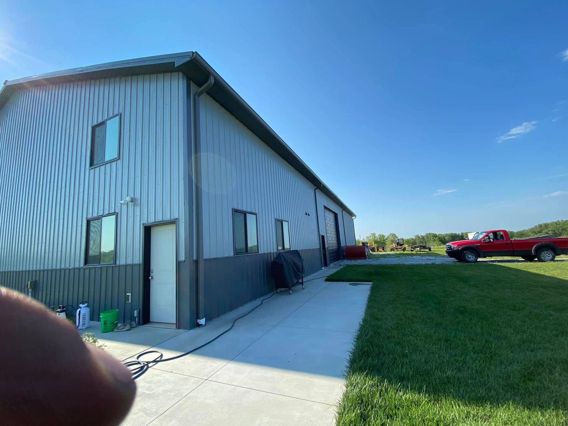 Gray industrial building beside a sidewalk and lawn under a blue sky, with a red pickup truck parked nearby