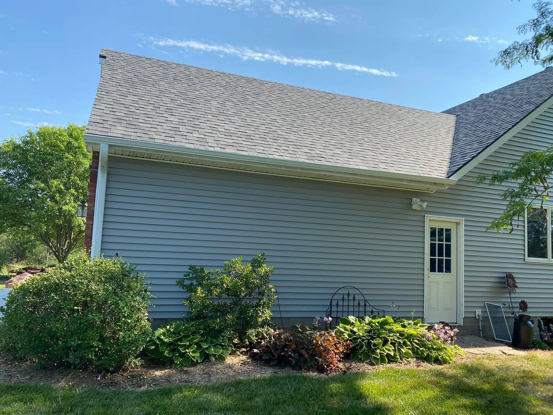 Suburban house exterior with gray siding, landscaped bushes, and a sunlit roof under a blue sky