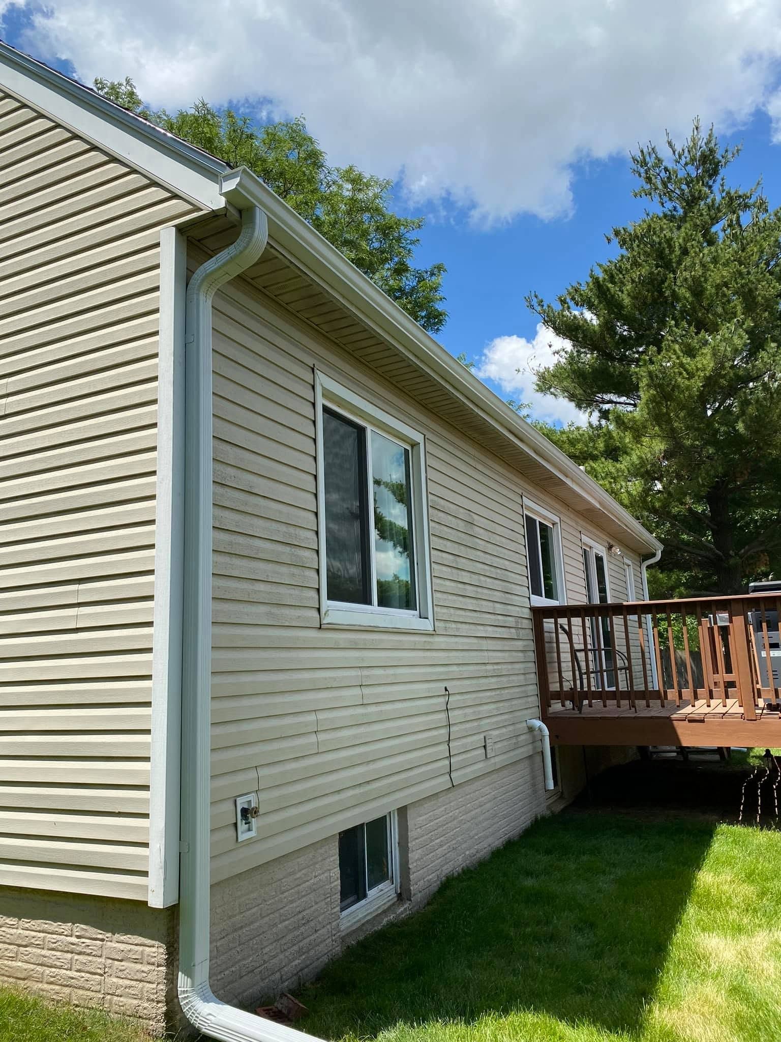 Side view of beige house with white trim, window, small deck, and green lawn under a partly cloudy sky