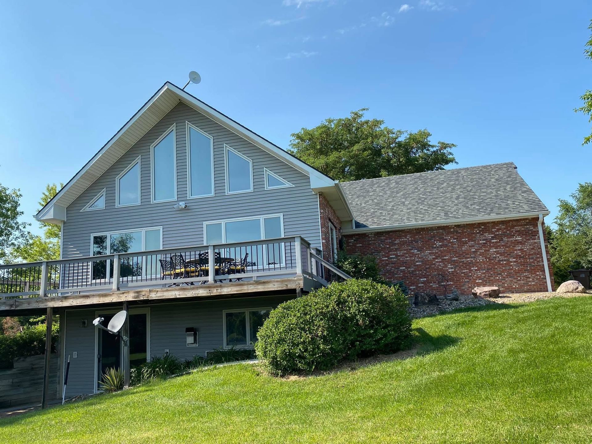 Two-story gray house with deck and brick side, set on a grassy hill under a blue sky