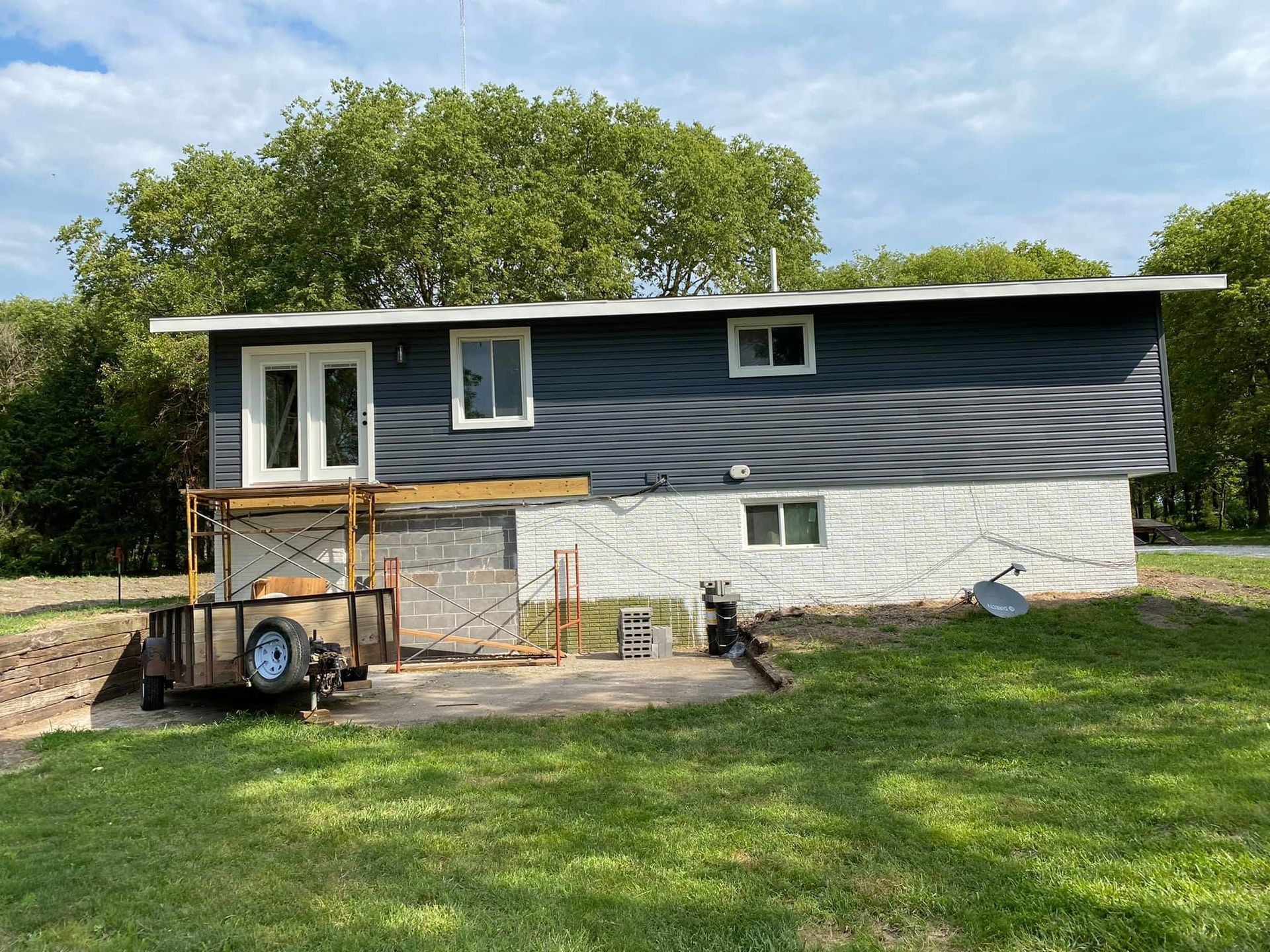 Small blue house with white lower siding, a porch, and yard equipment in front under a sunny sky