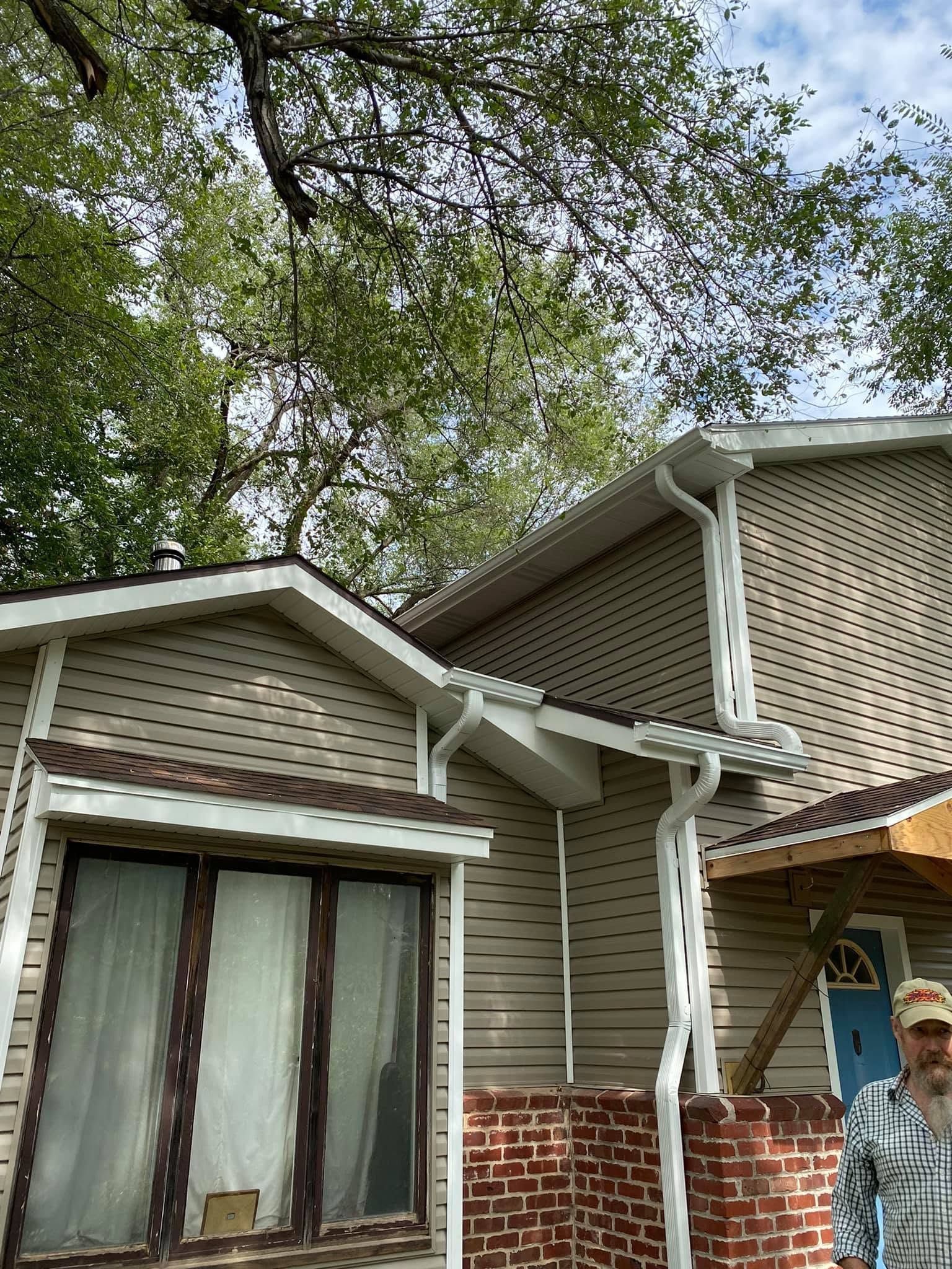 Two-story house exterior with beige siding, brick base, and large trees overhead