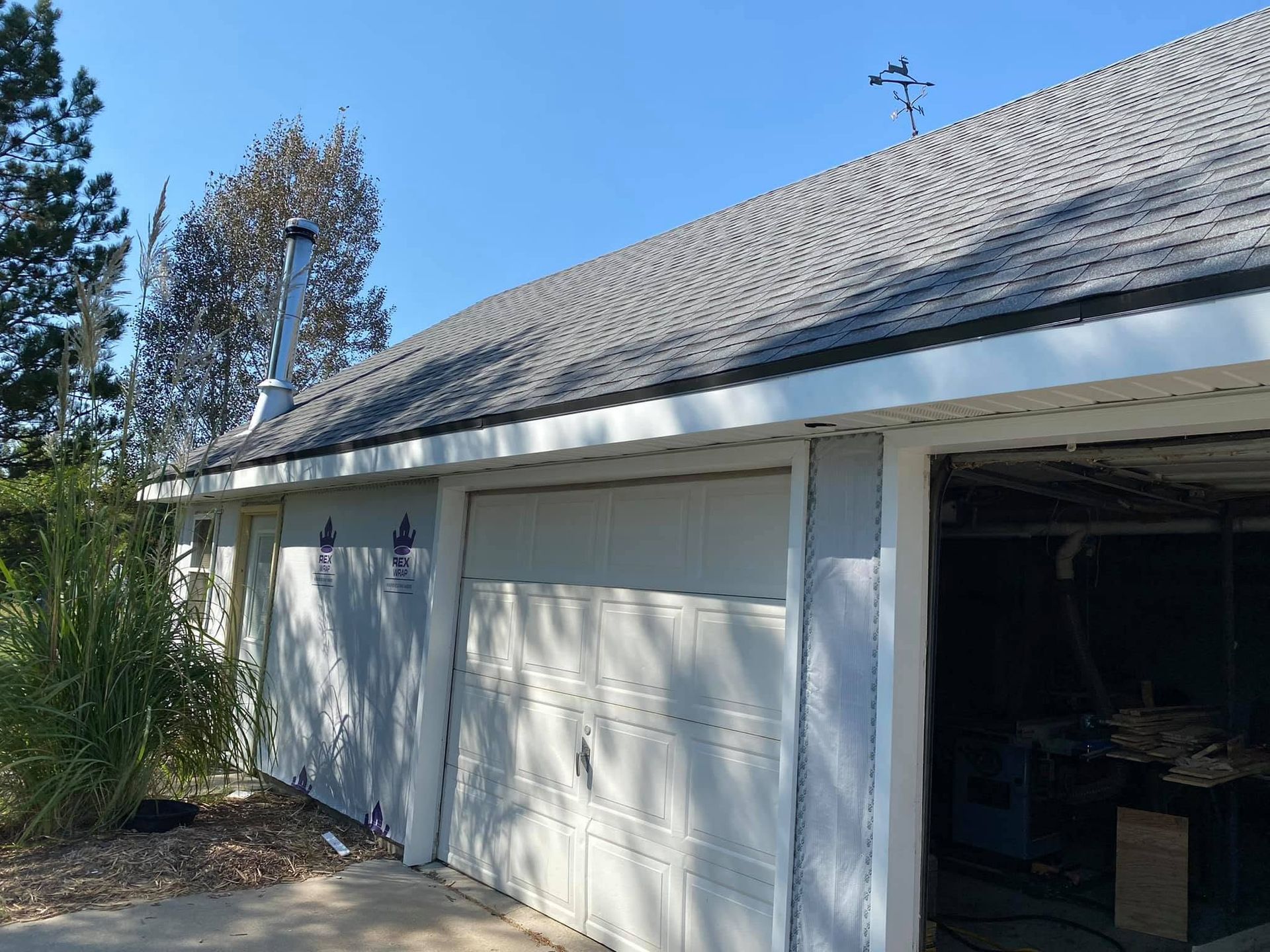White garage with open bay and closed doors, viewed from a driveway beside trees and bushes.
