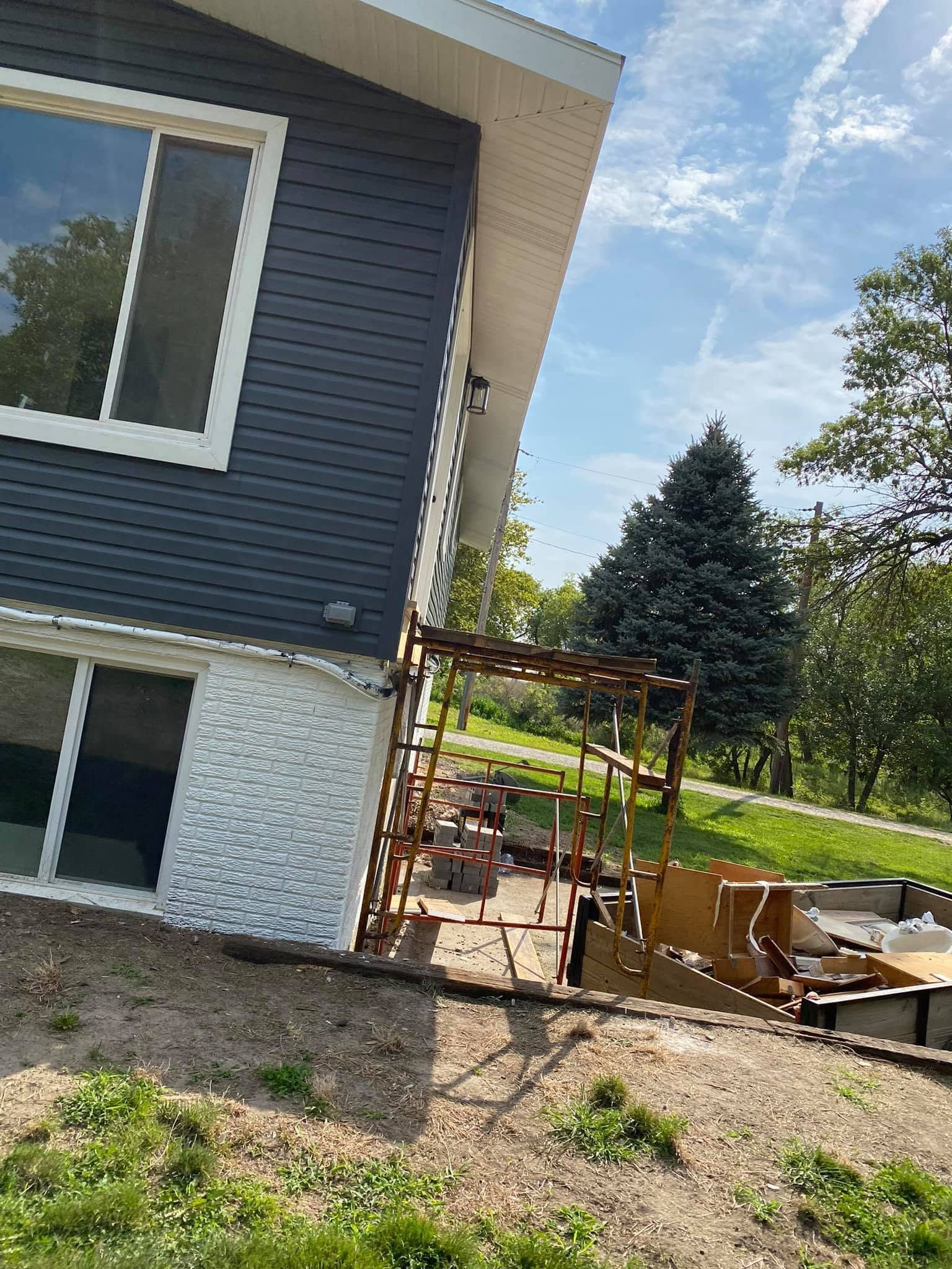 Side of a blue house with a window, showing a damaged deck and excavated ground in a yard.