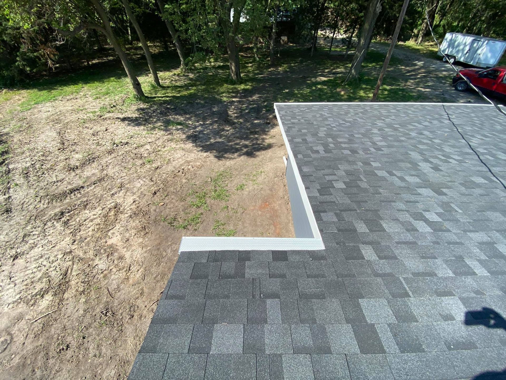 Fresh gray shingle roof edge beside a dirt yard with trees in the background