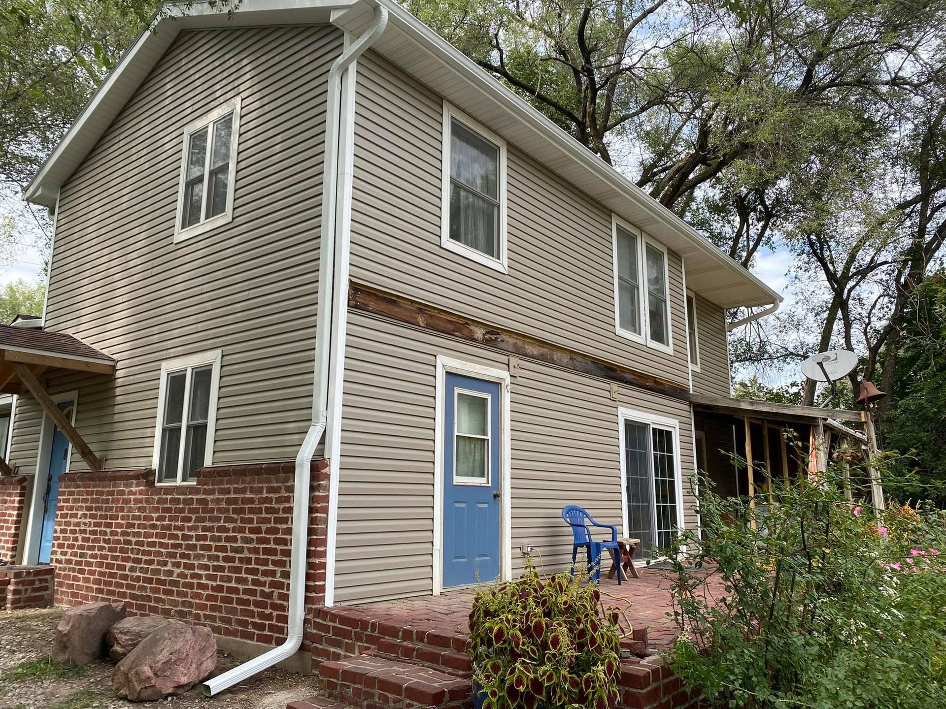 Two-story house with tan siding and red brick, blue front door, and a small porch surrounded by trees and shrubs.