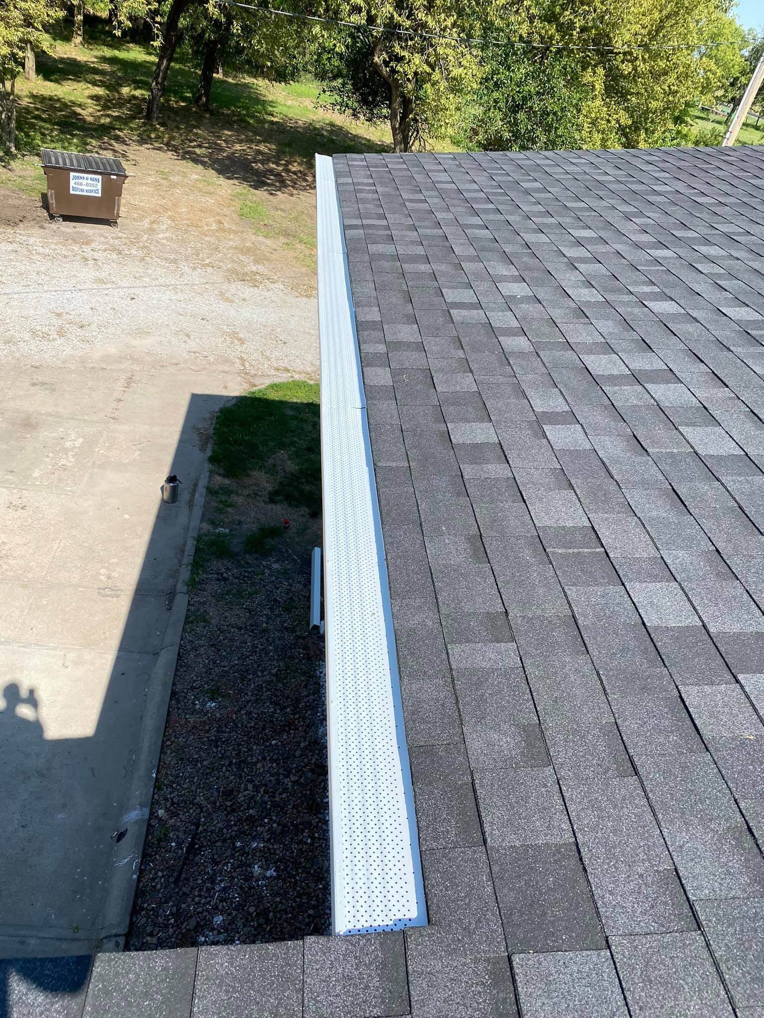 Aerial view of a gray shingled roof with a white gutter beside a driveway and gravel strip.