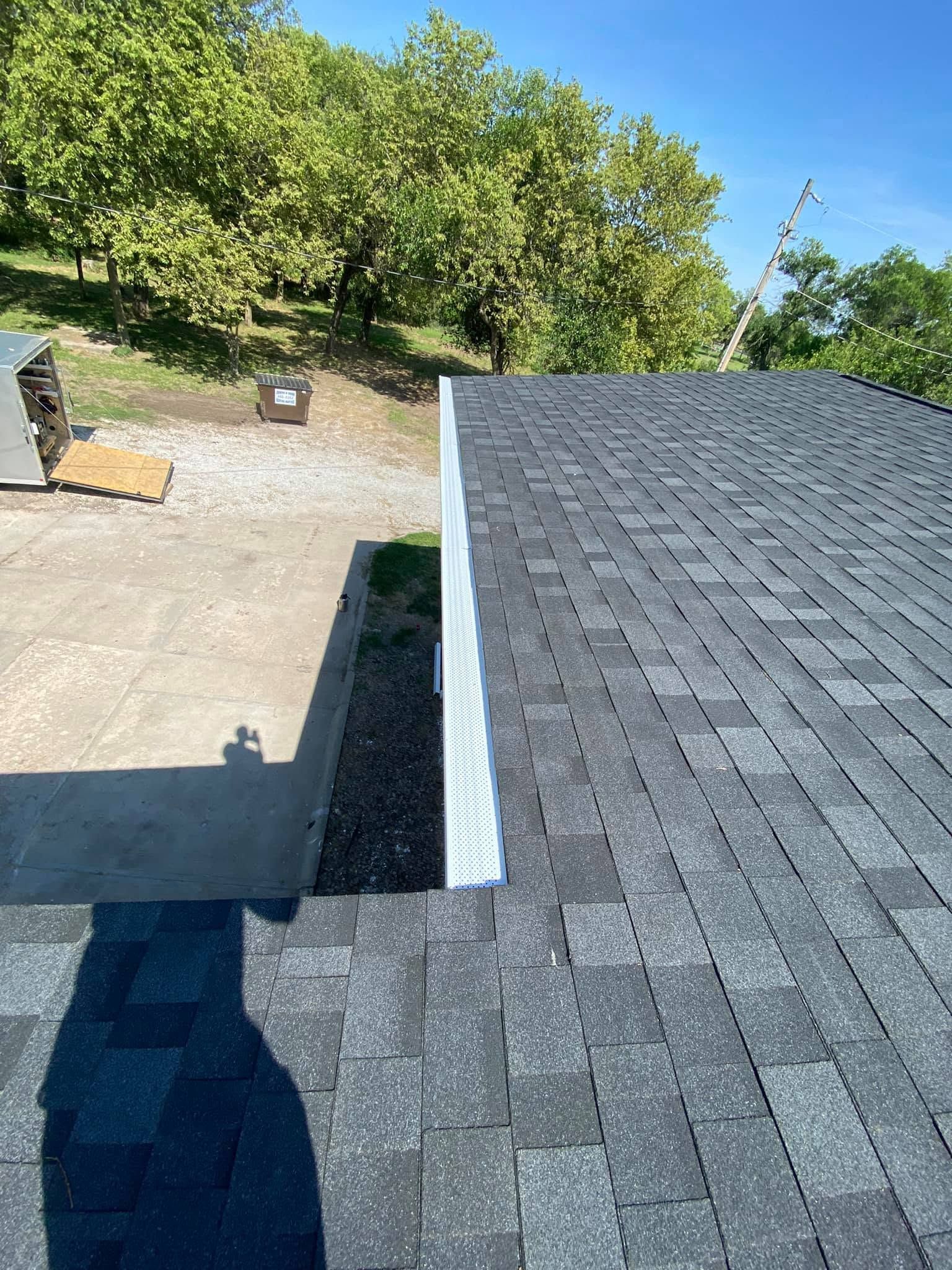 Roof shingles beside a gravel driveway and green trees under a clear blue sky