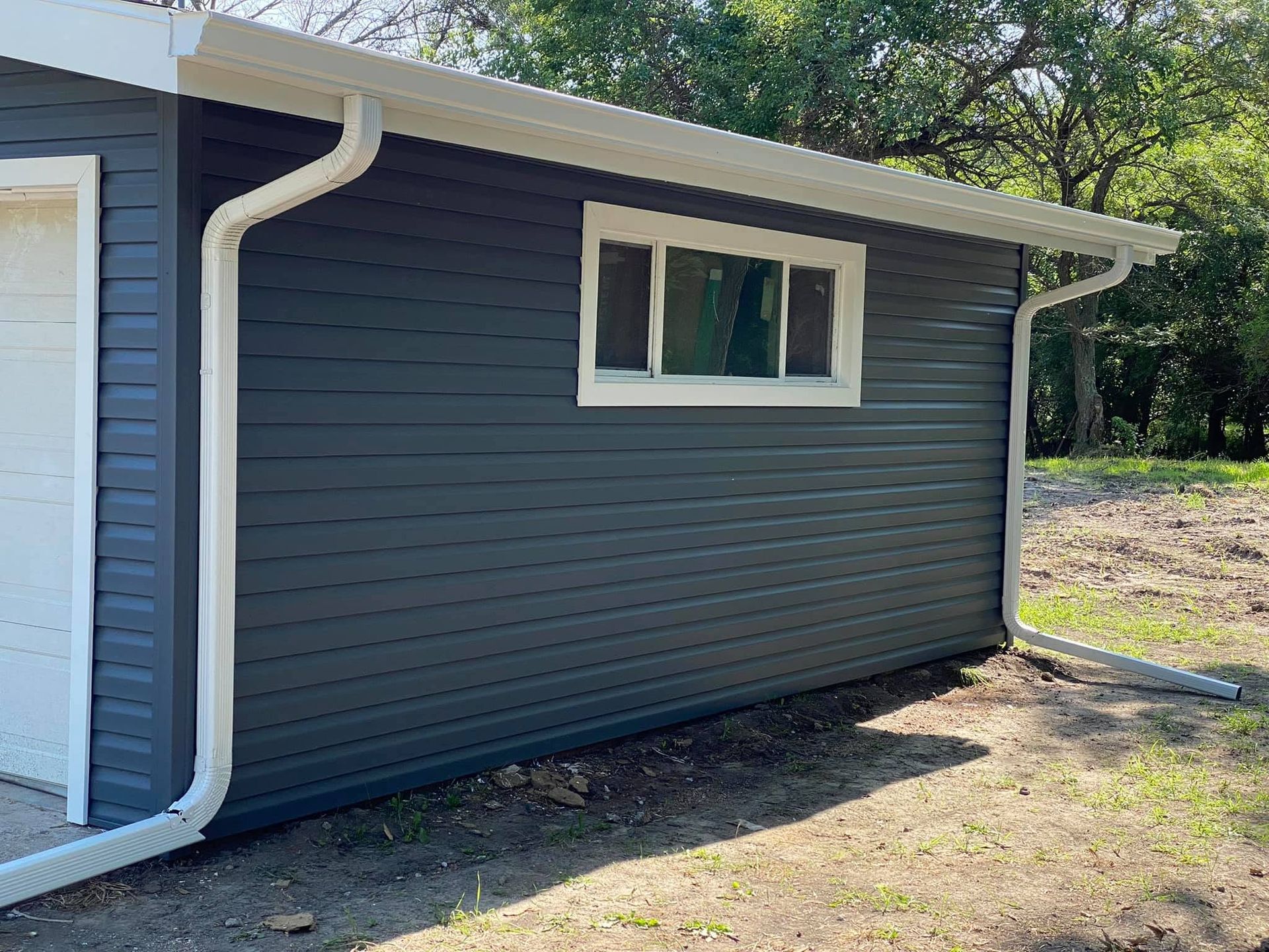 Gray house exterior with white trim, small window, and white downspout beside a dirt yard