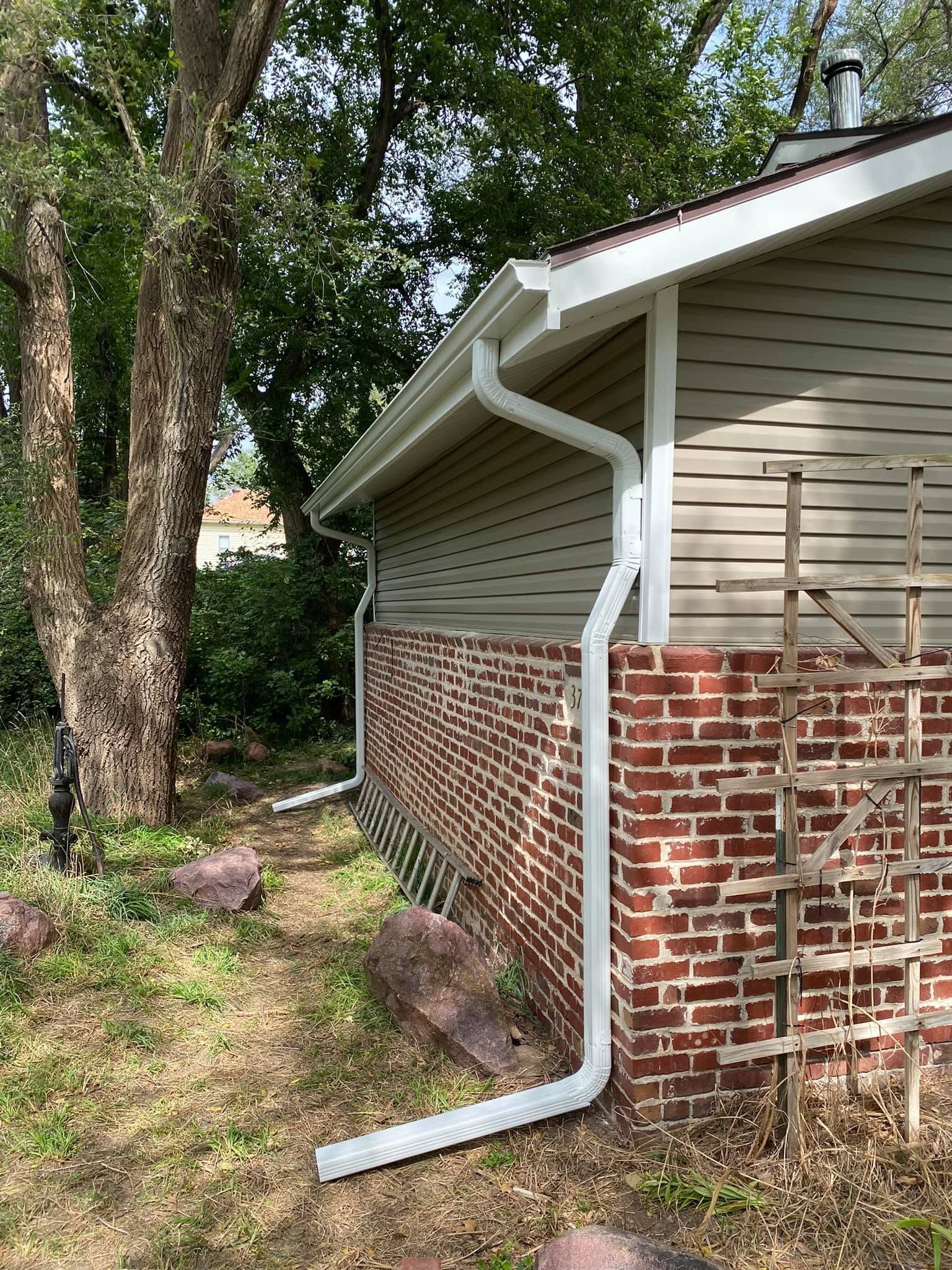 White downspout on a red brick and beige house exterior beside a tree and yard greenery