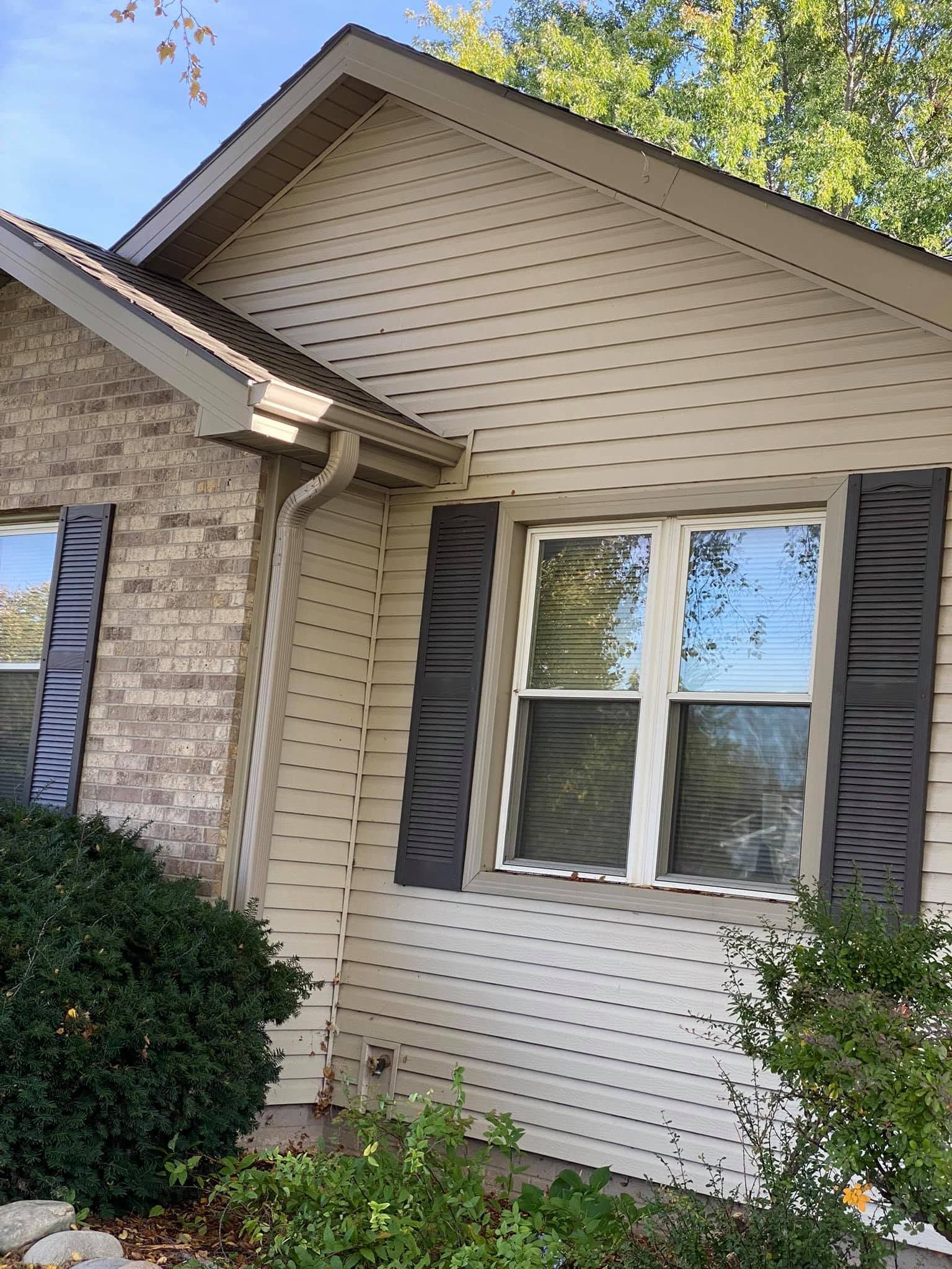 House exterior with beige siding, dark shutters, a front window, and shrubs under a gabled roof.