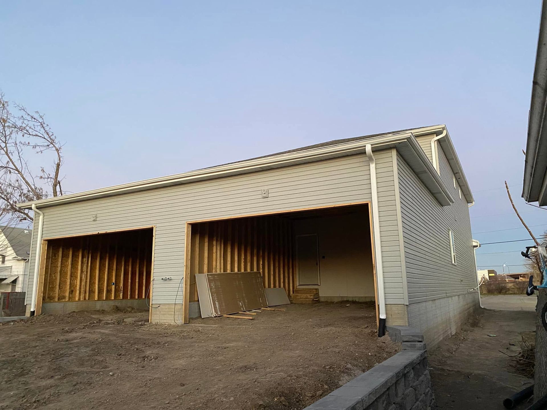 Partially built two-car garage with white siding and open bays on a dirt lot