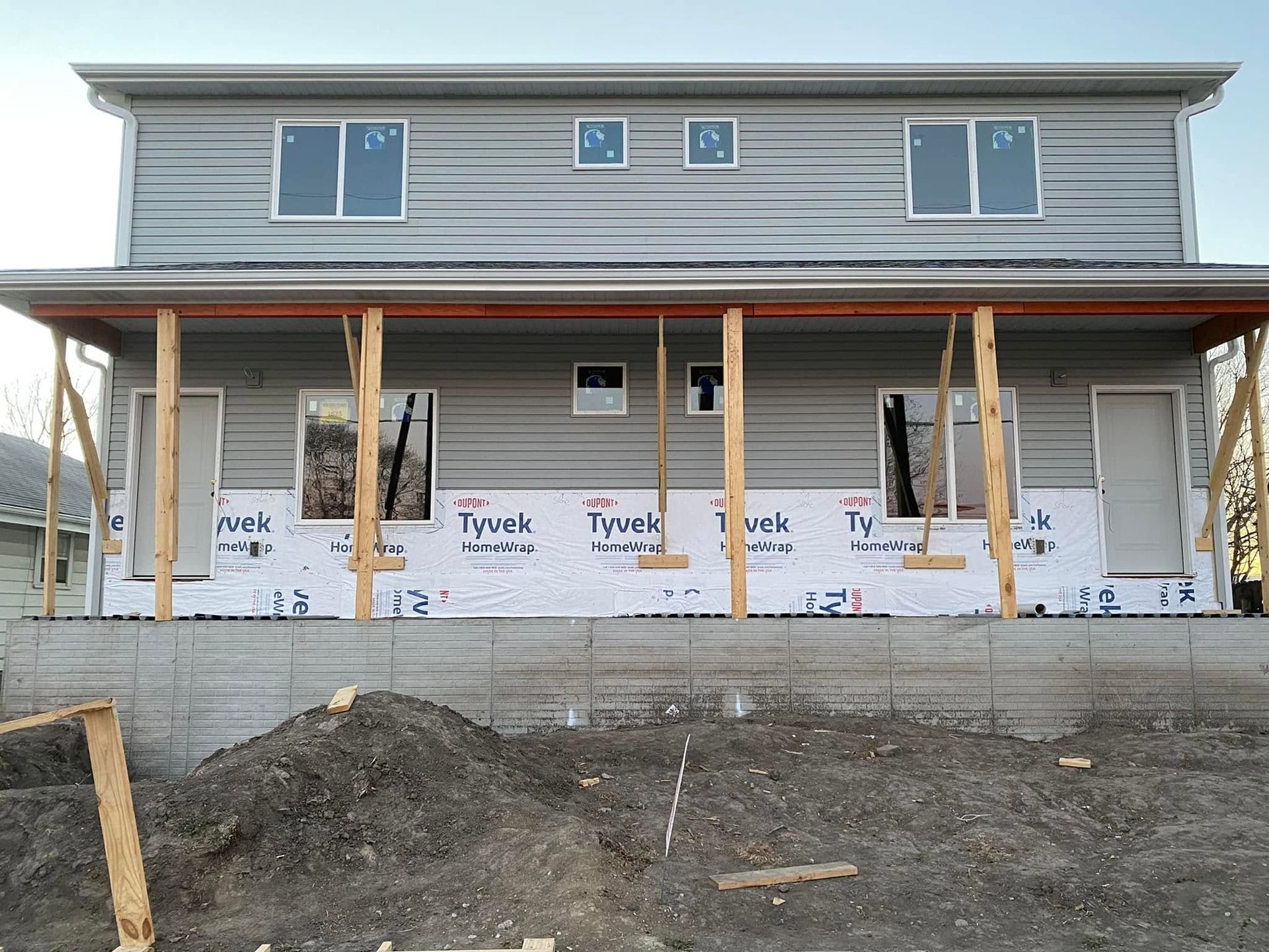 Front of a two-story house under construction with exposed framing, plywood sheathing, and a dirt foreground