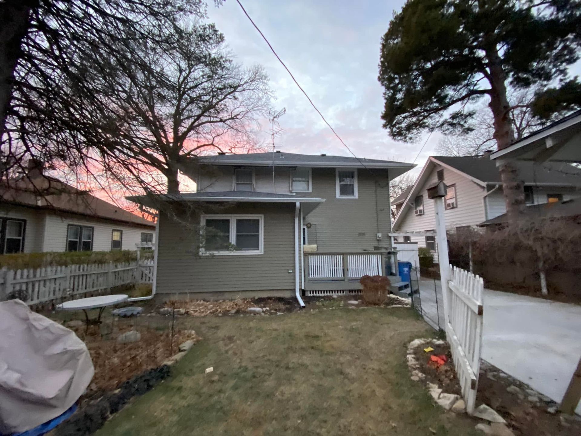 Backyard view of a gray two-story house at dusk with a fenced yard and pink sky
