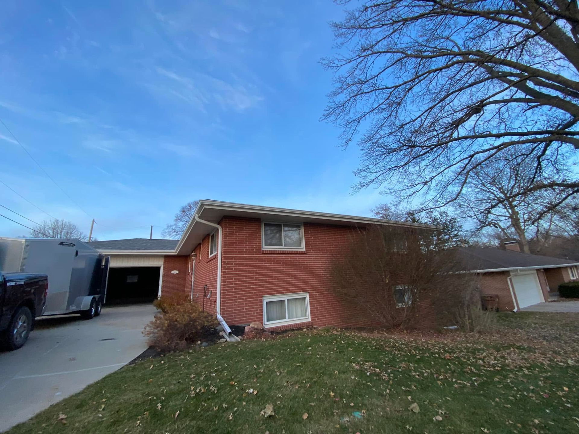 Red brick house with driveway, parked trailer, and grassy yard under a blue sky.