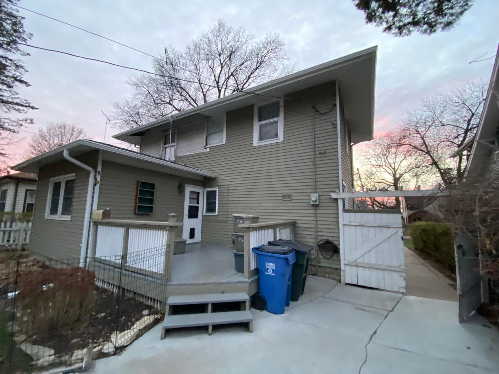 Two-story beige house with a small porch, driveway, blue recycling bin, and fenced side yard at dusk