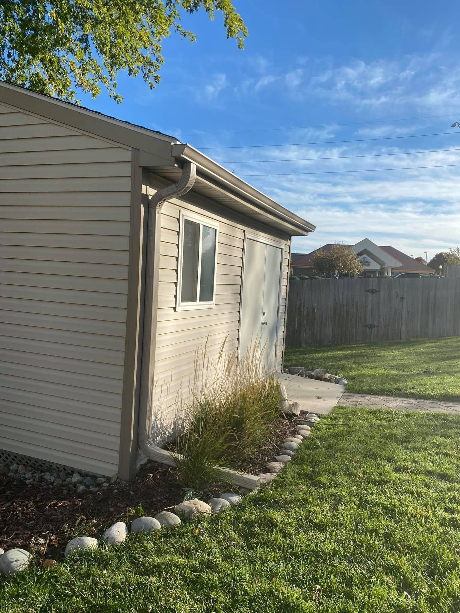 Backyard side view of a beige house with white trim, stone edging, grass, fence, and blue sky