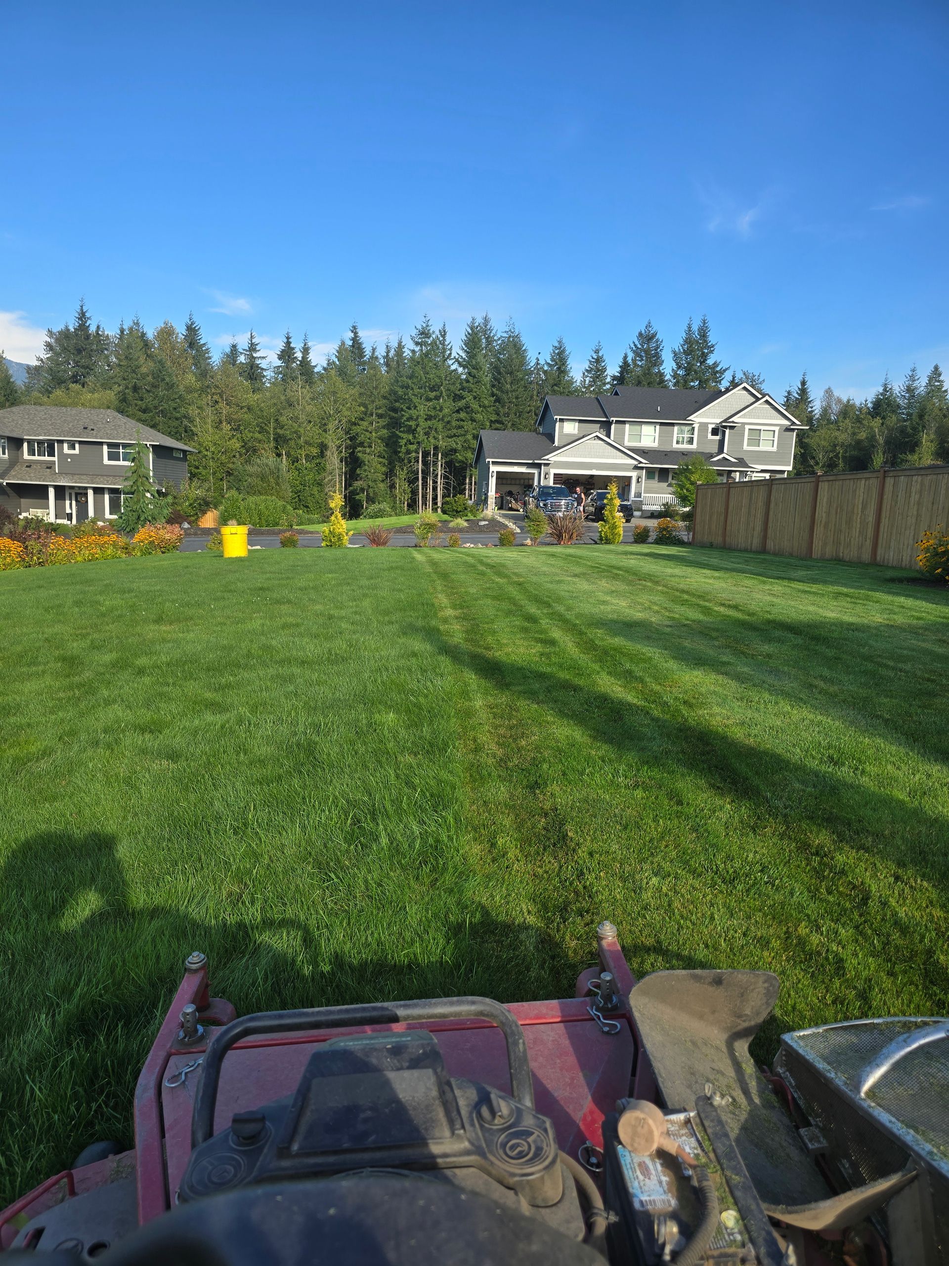 Wide backyard with green lawn, trees, houses, and a lawn mower at the bottom edge under a blue sky