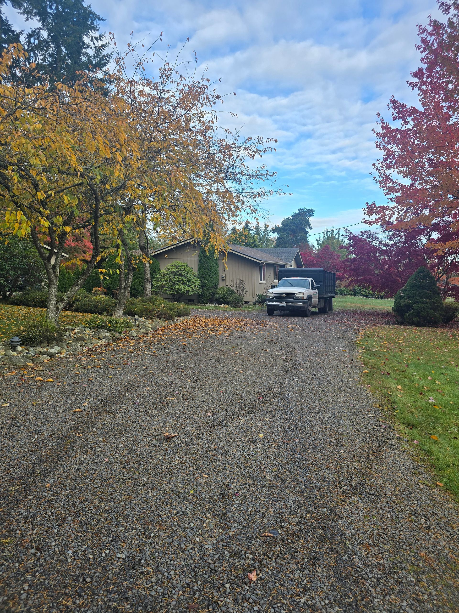 Gravel driveway with a parked truck in front of a house, surrounded by trees with autumn foliage. Cloudy sky.