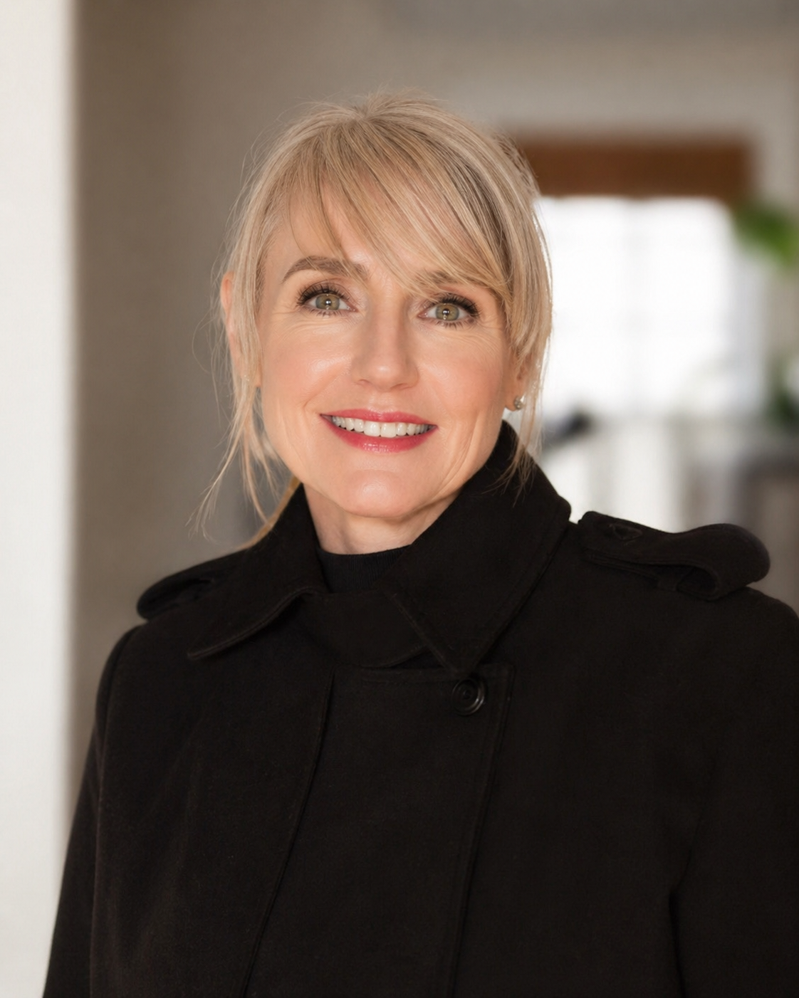 Smiling blonde woman in a black coat against a softly blurred indoor background