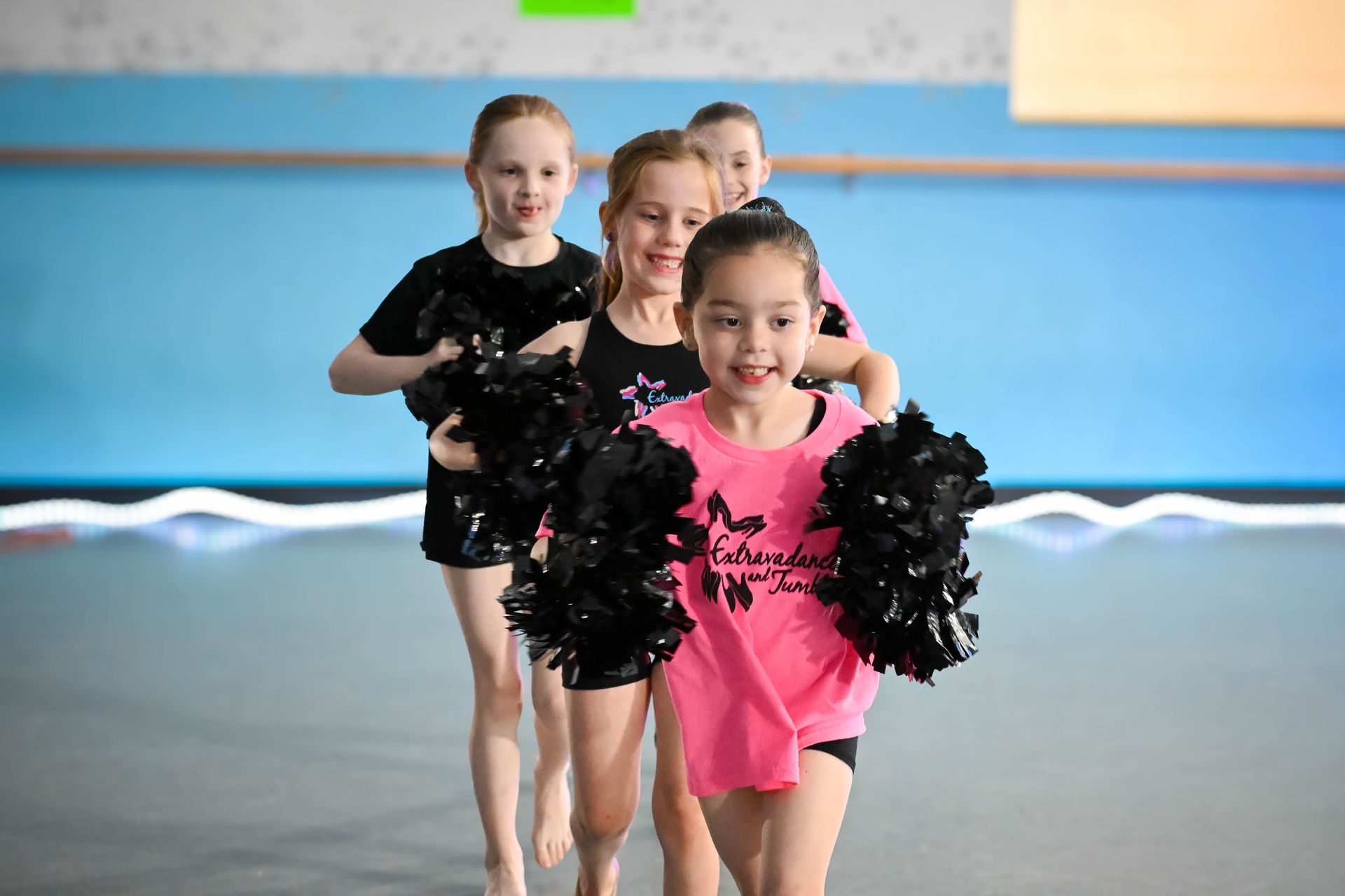Three cheerleaders in black and pink uniforms pose on an indoor rink, smiling and holding pom-poms.