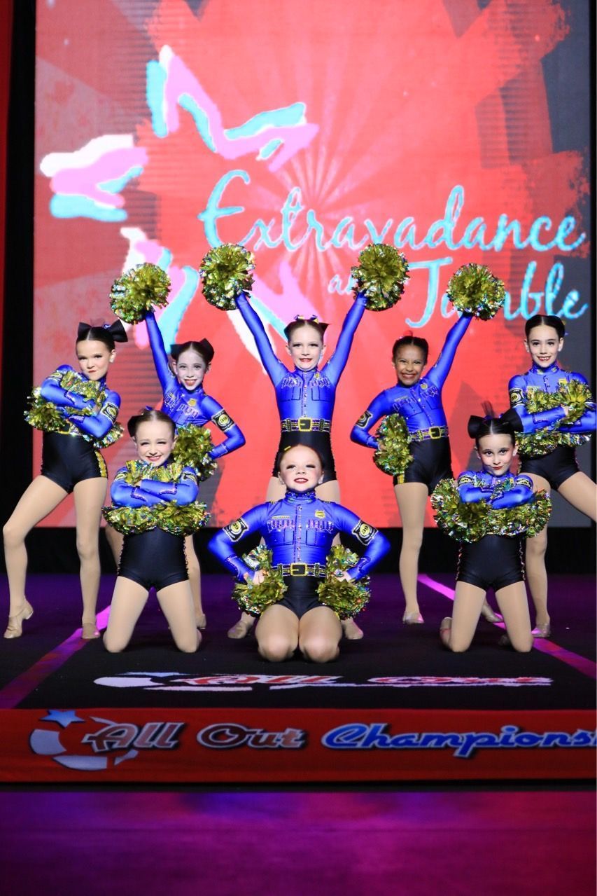 Youth dance team in blue and black costumes with green pom-poms posing on stage in front of a red backdrop.