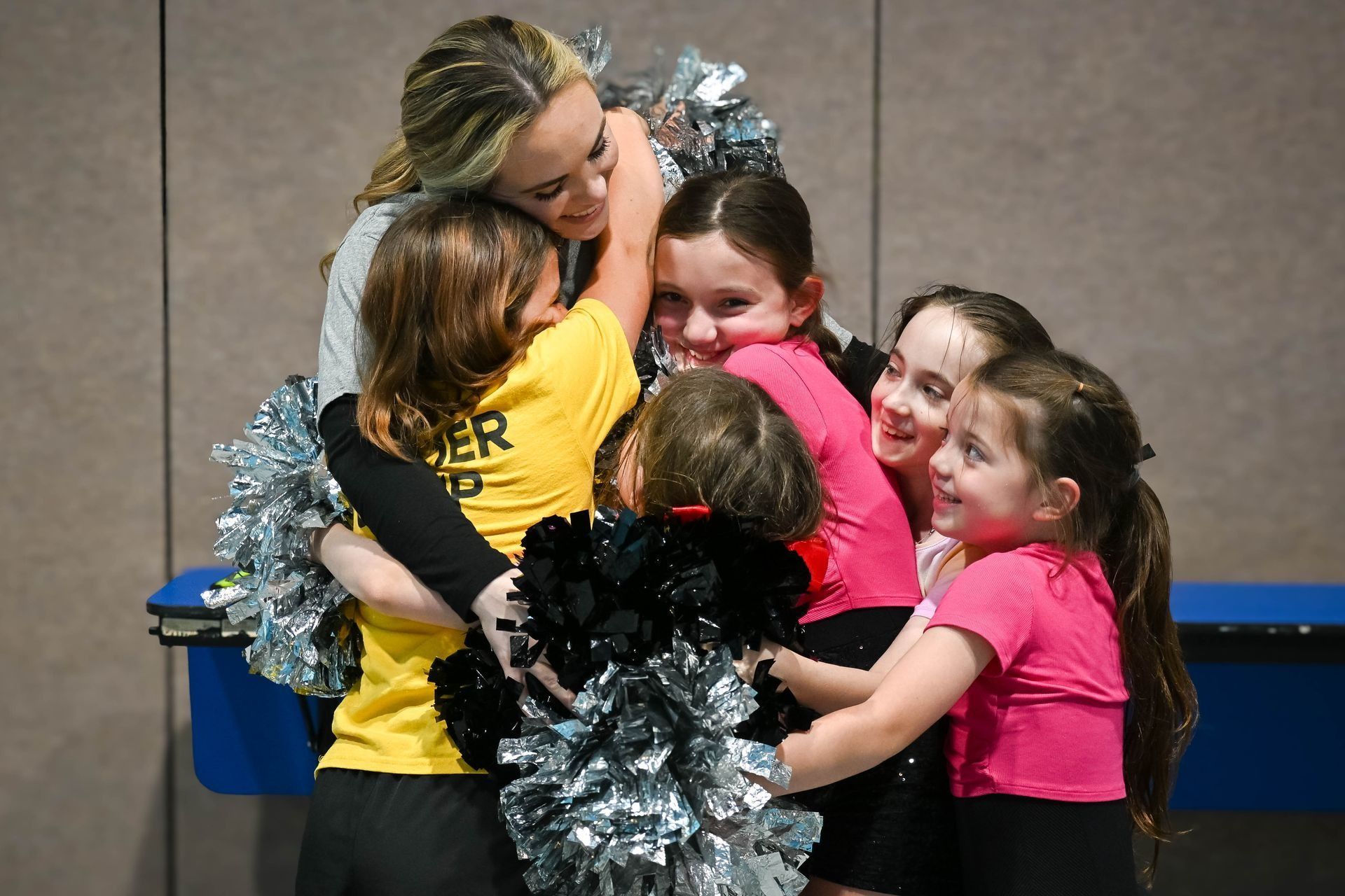 Four children in cheer uniforms hug in a group, holding silver pom-poms, with an adult coach leaning over them.