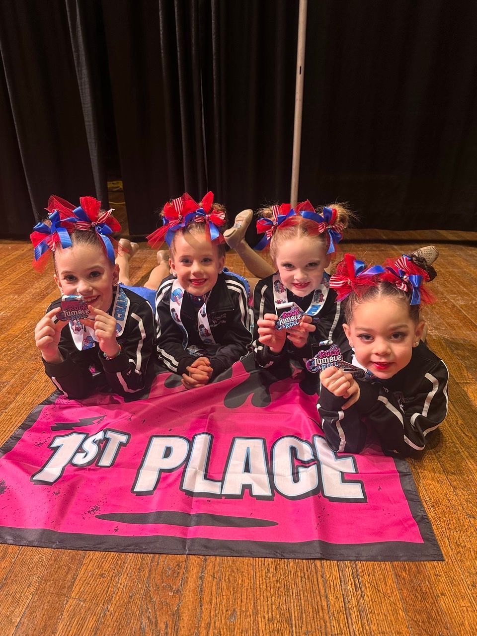 Four young dancers kneel on a pink 1st Place mat, wearing black outfits and red-blue bows.