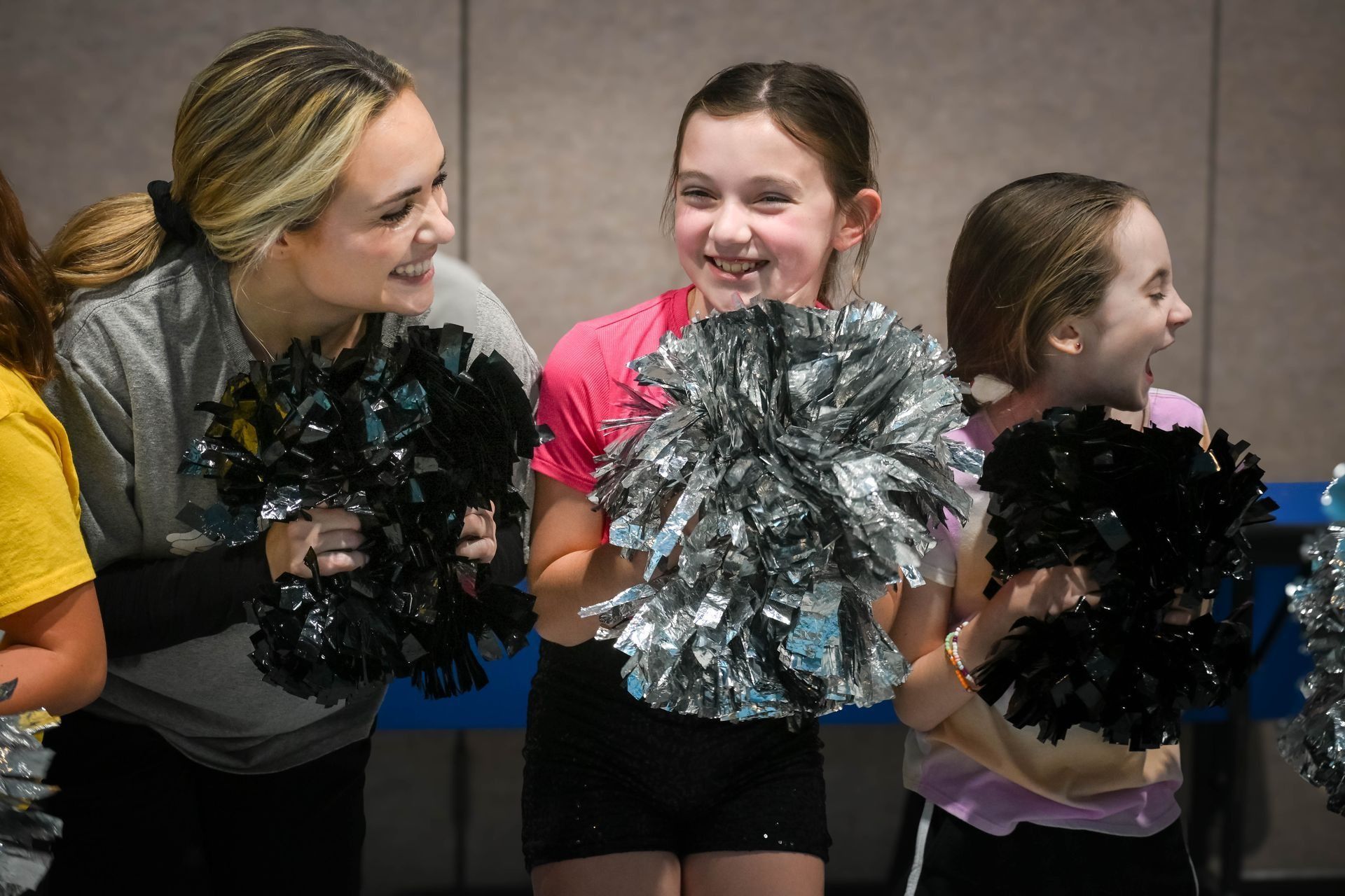 Three cheerleaders holding silver and black pom-poms, smiling together indoors