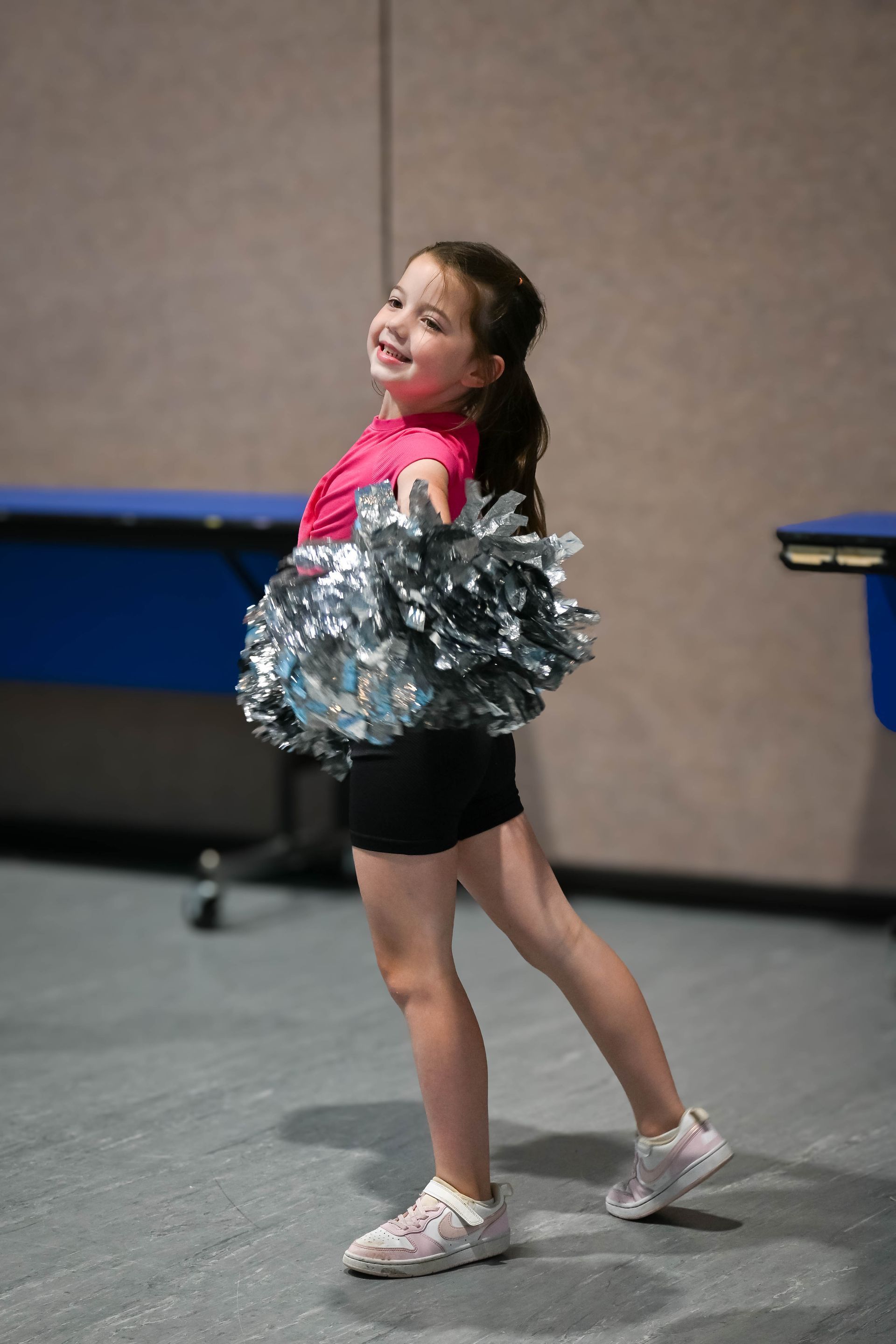 Young cheerleader in pink and black outfit posing with silver pom-poms in a gym