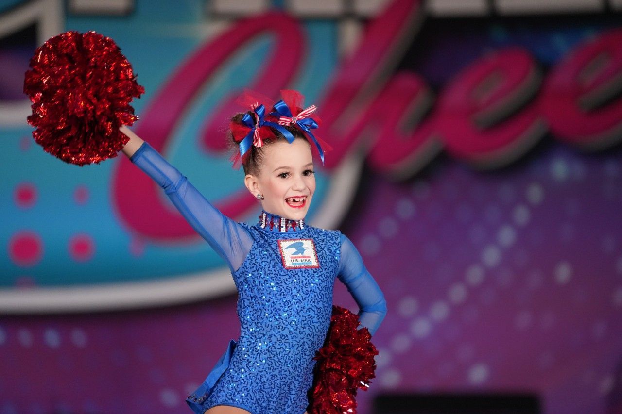 Young cheerleader in blue uniform with red pom-poms performing onstage, smiling with one arm raised.