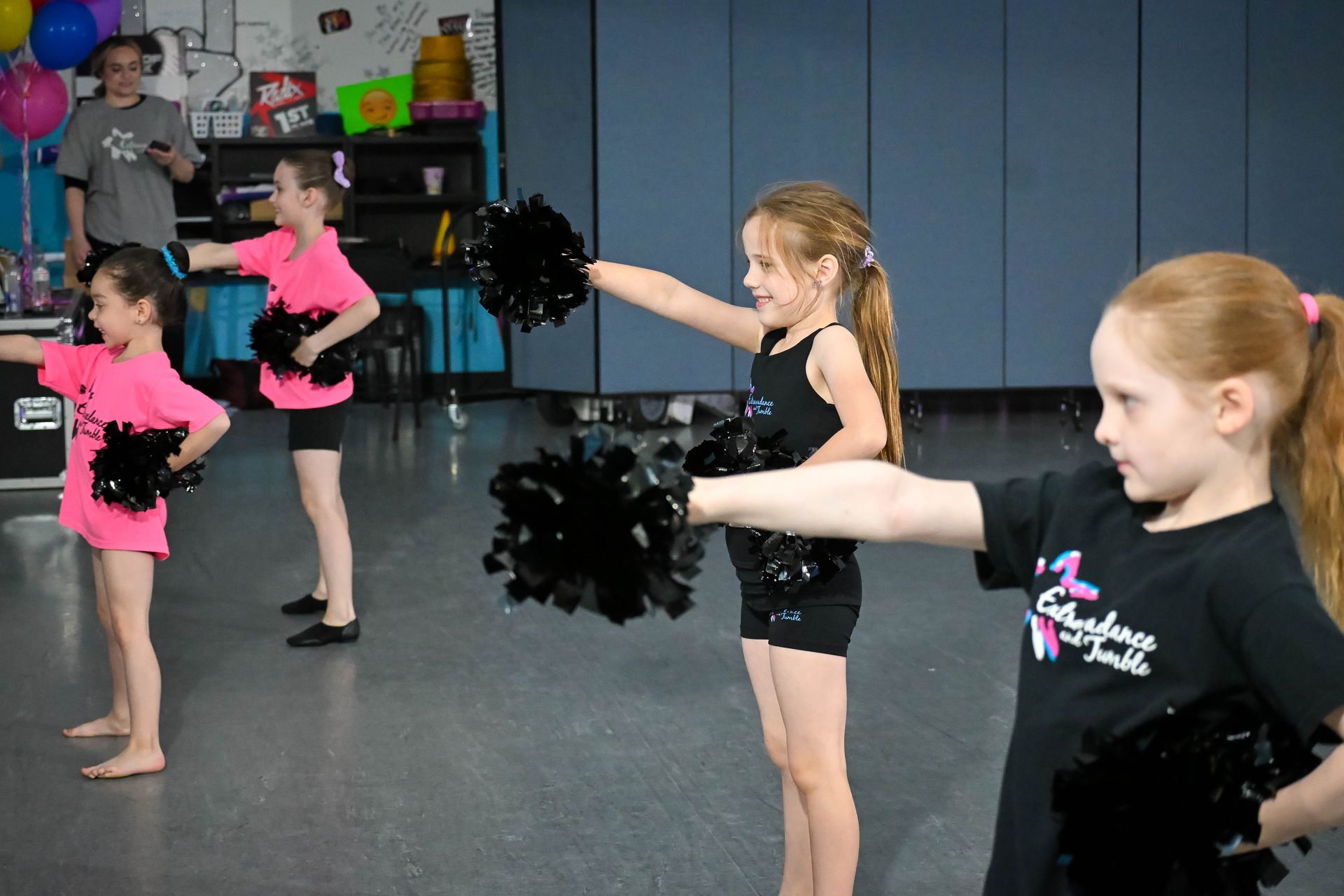Children in black and pink dance outfits practicing pom-poms in a studio