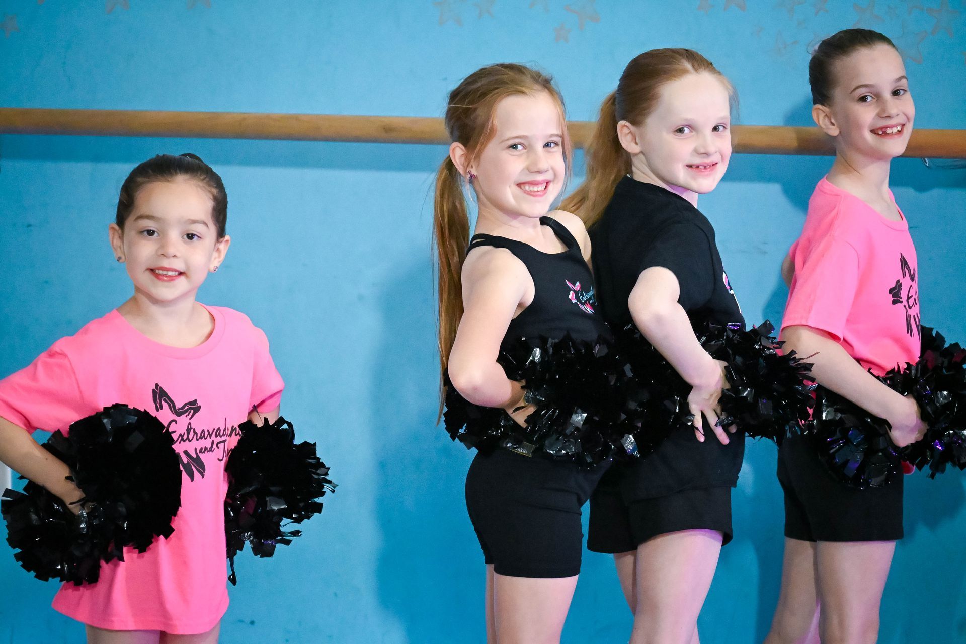 Four young dancers in pink and black cheer outfits pose with pom-poms against a blue wall.
