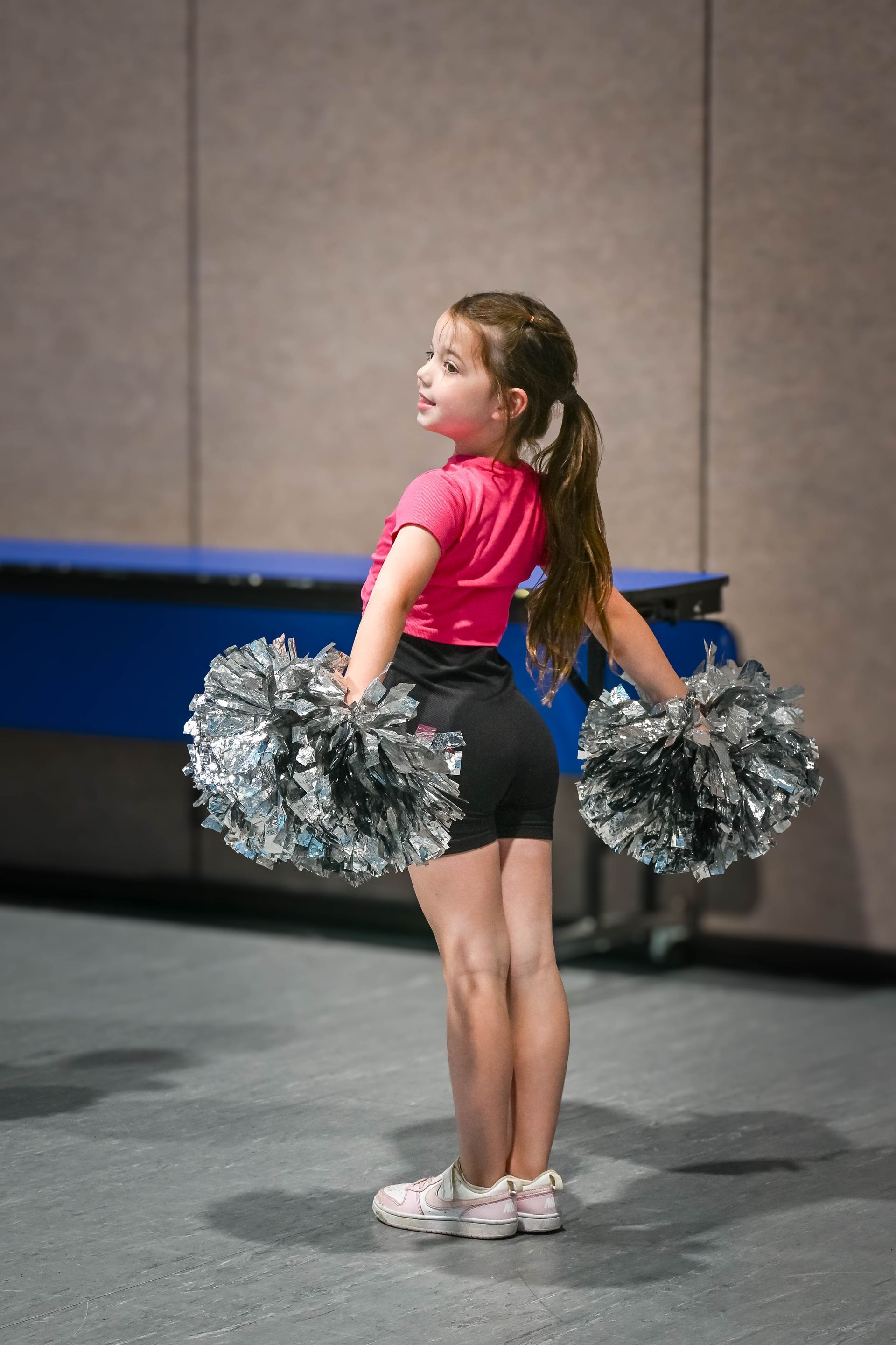 Young cheerleader in a pink top and black shorts holding silver pom-poms on a stage