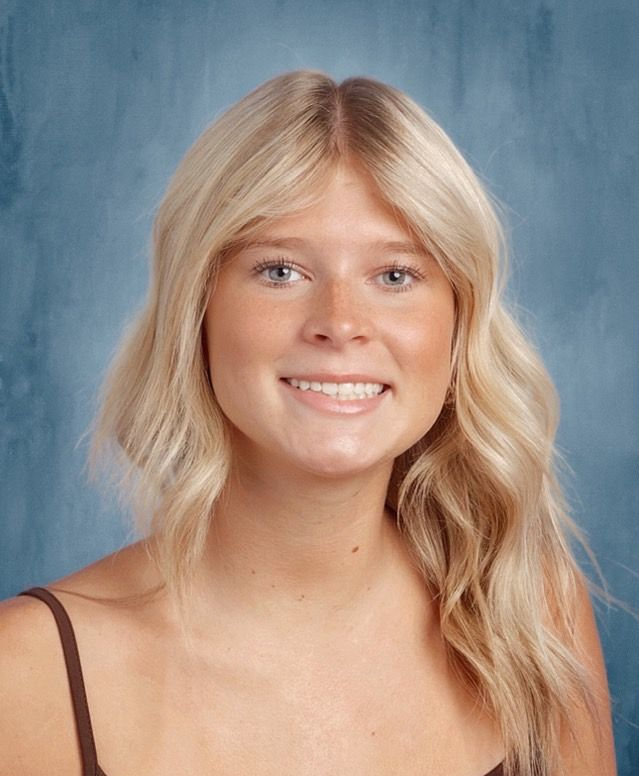 Smiling blonde person in a black top against a blue studio backdrop