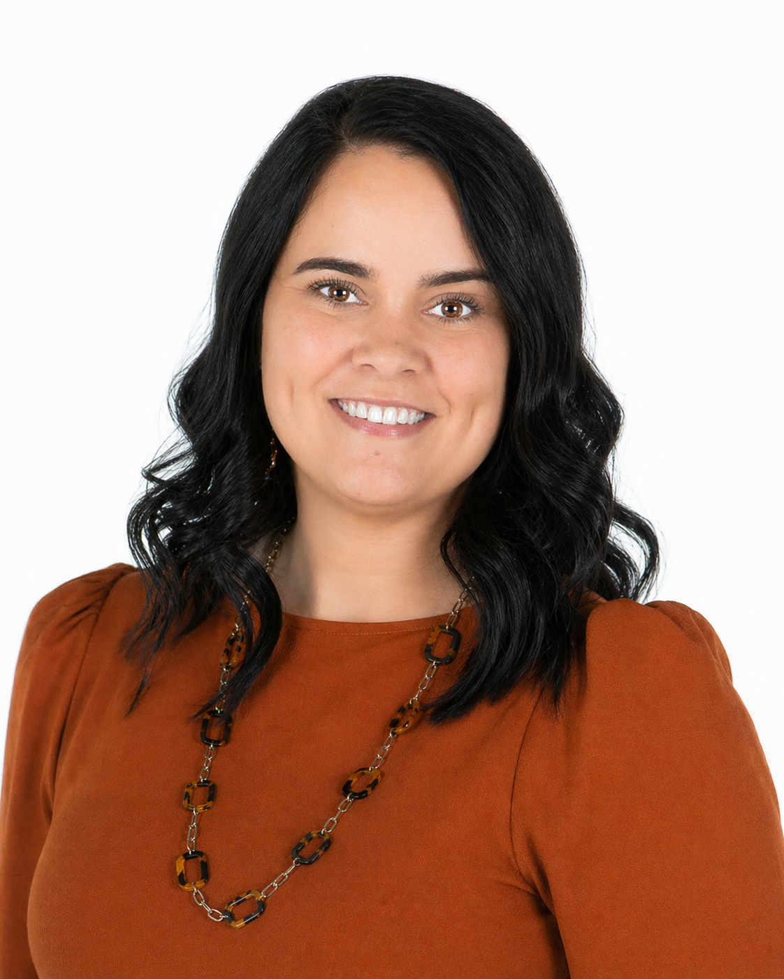 Smiling woman with black hair wearing an orange top and beaded necklace against a white background