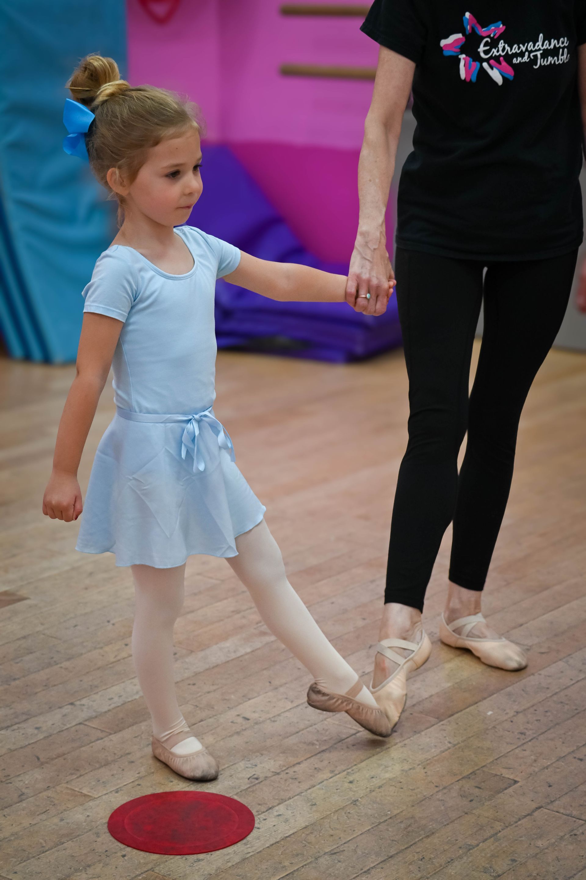 Young ballet dancer in a blue dress holding an instructor’s hand in a dance studio.