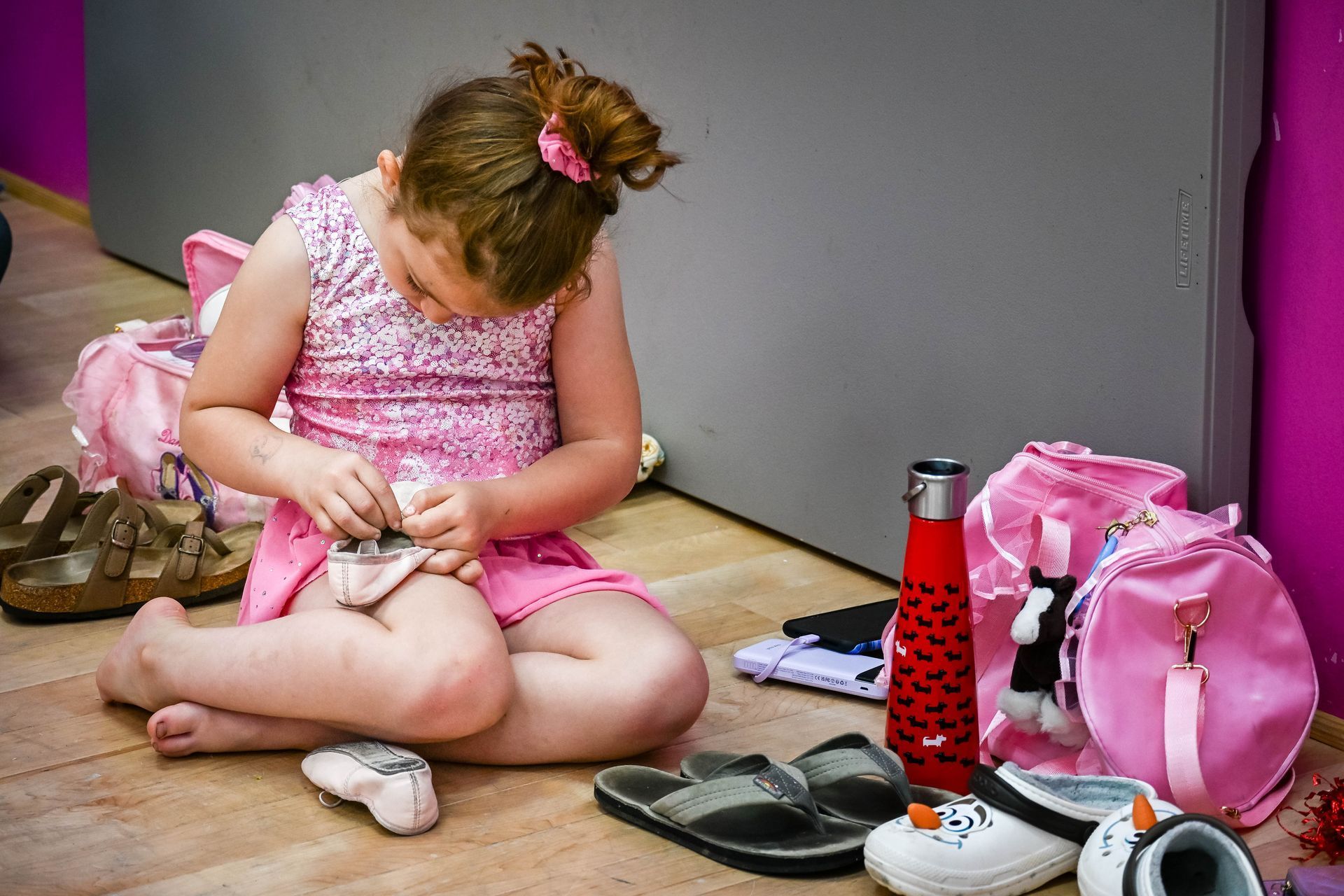 Child sitting on the floor packing a pink backpack beside shoes and a red water bottle.