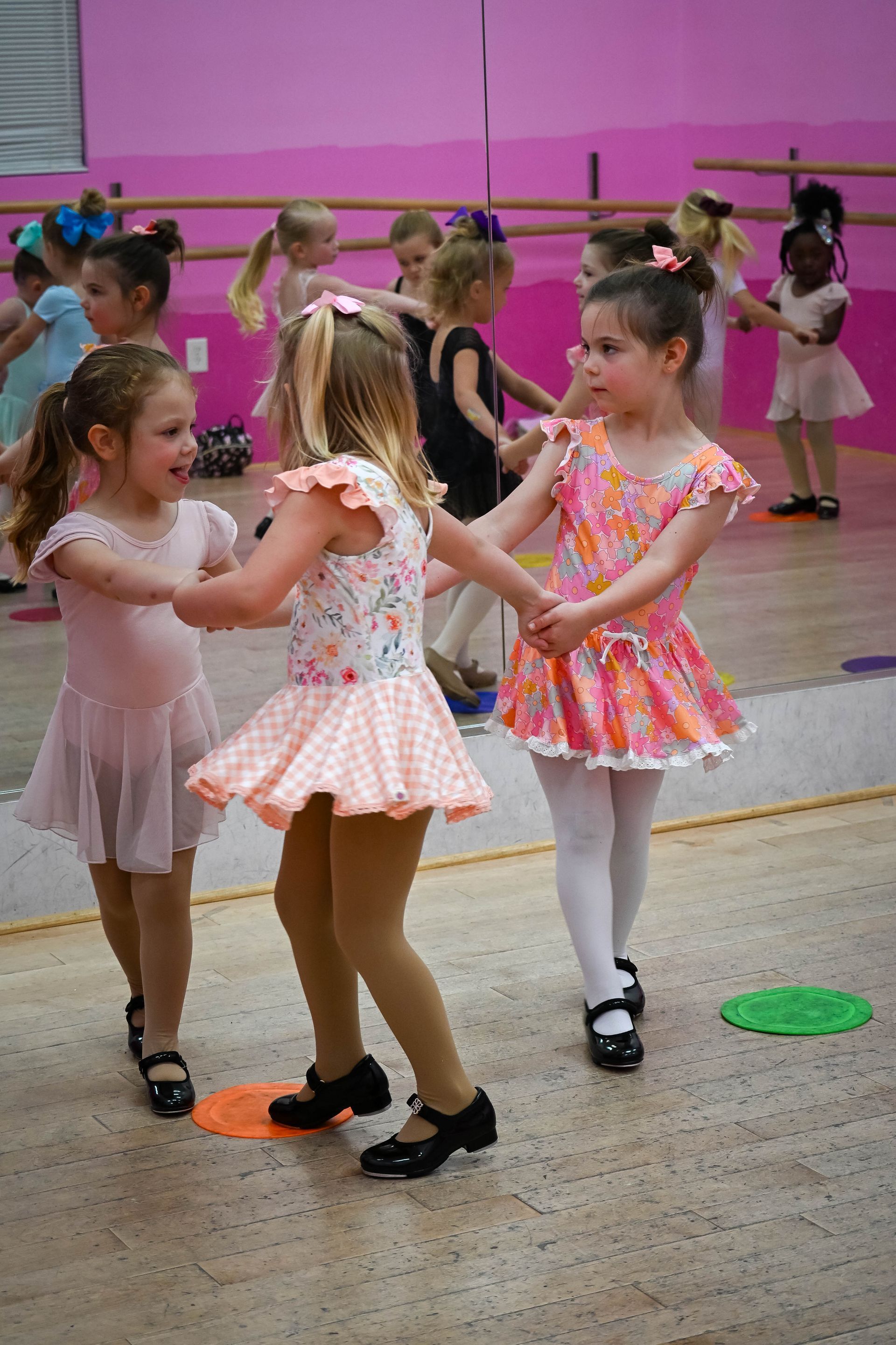 Children in pastel dresses holding hands and dancing in a studio with a pink wall.
