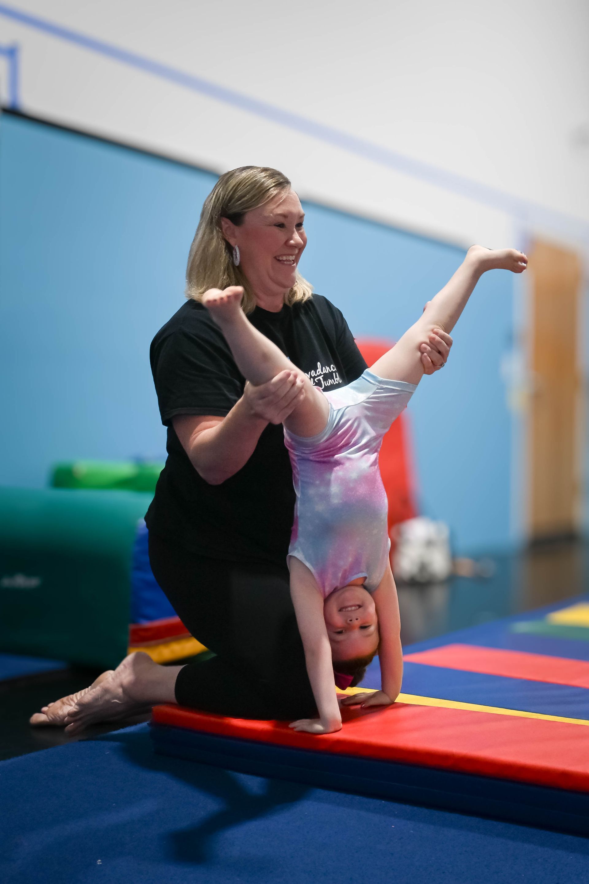 Woman assisting a child doing a handstand on a red mat in a gymnasium.