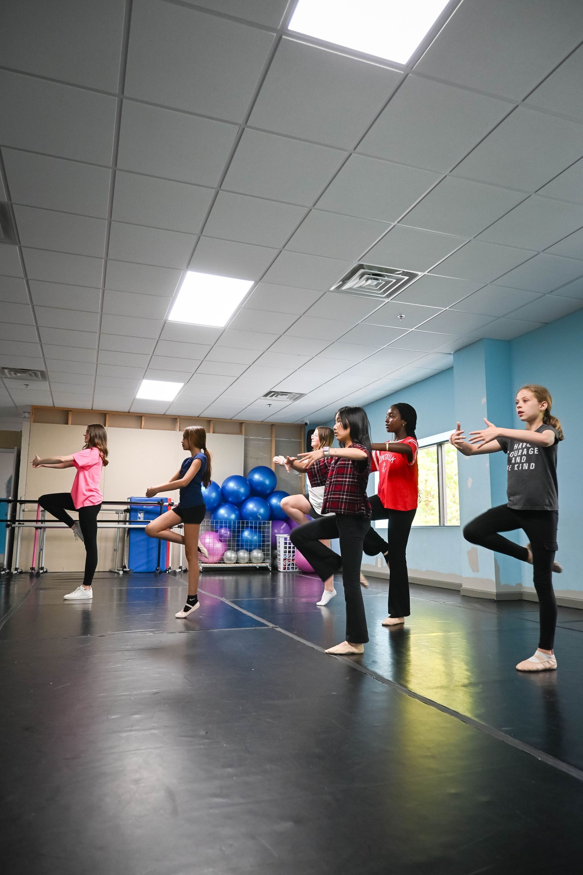 People balancing in a dance studio, practicing poses on one leg with arms extended.