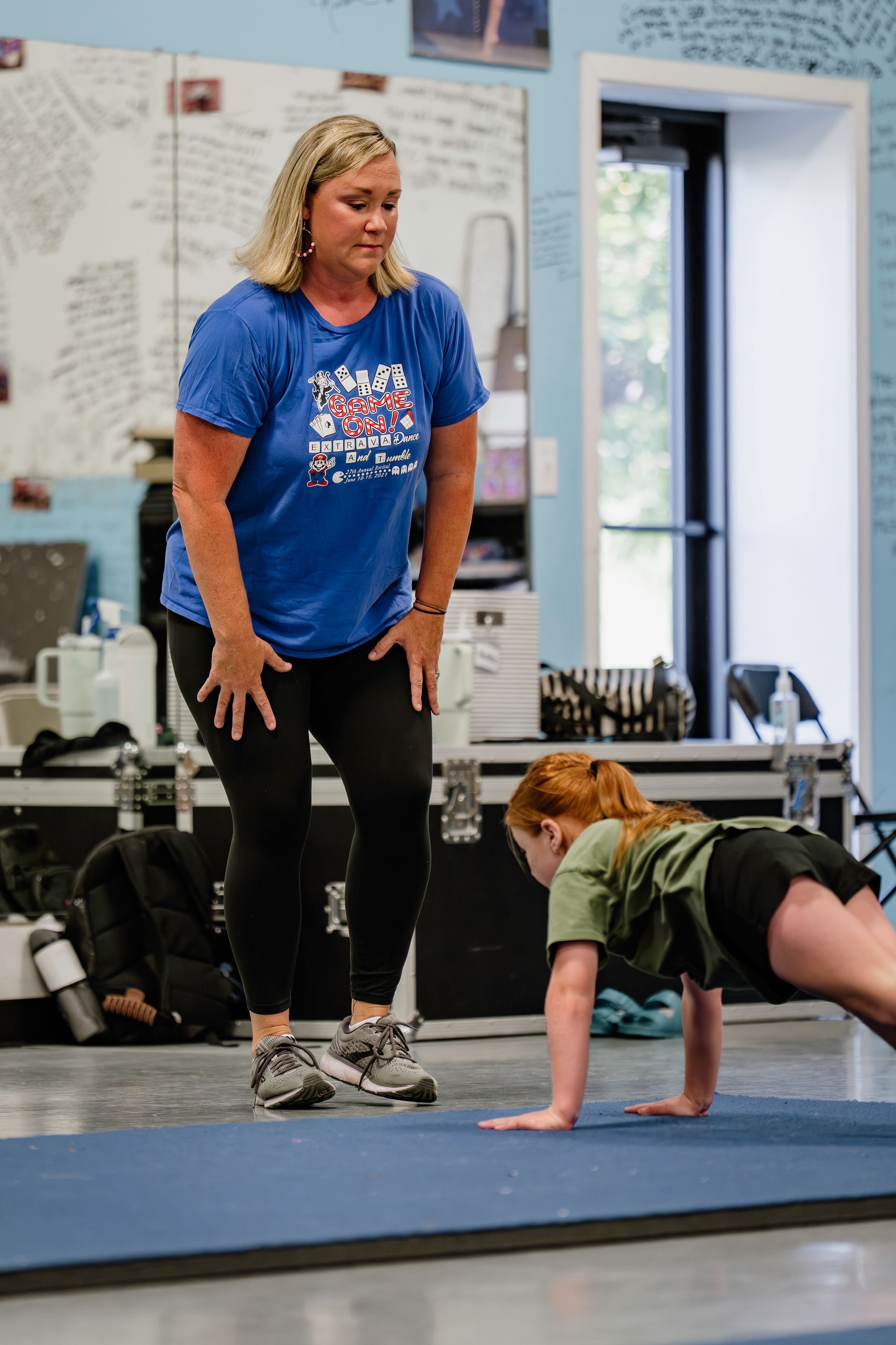 Coach and child doing floor exercises in a gym, with the coach standing nearby.
