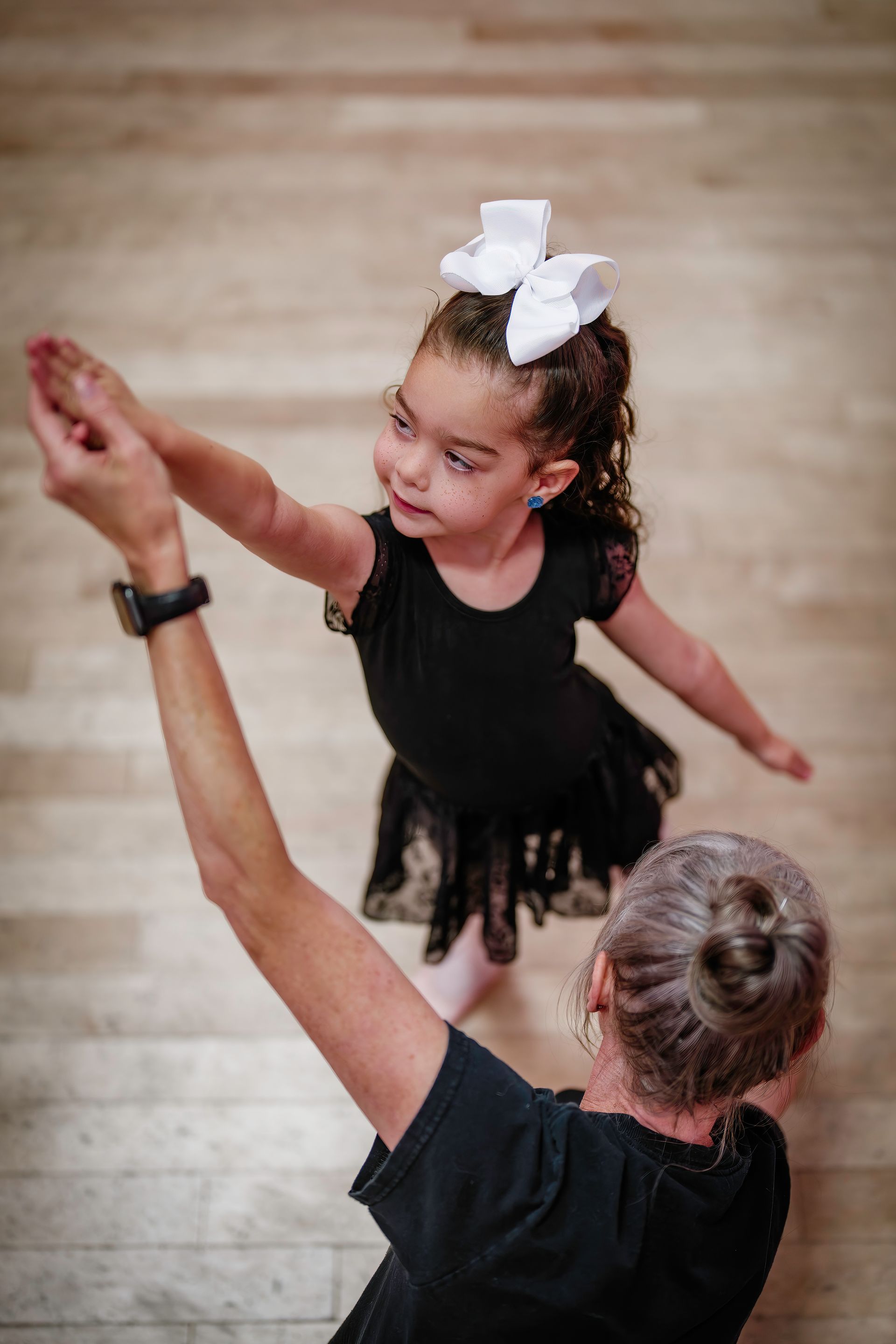 Dance instructor guiding a young dancer in a black dress with a large white bow on a wooden floor