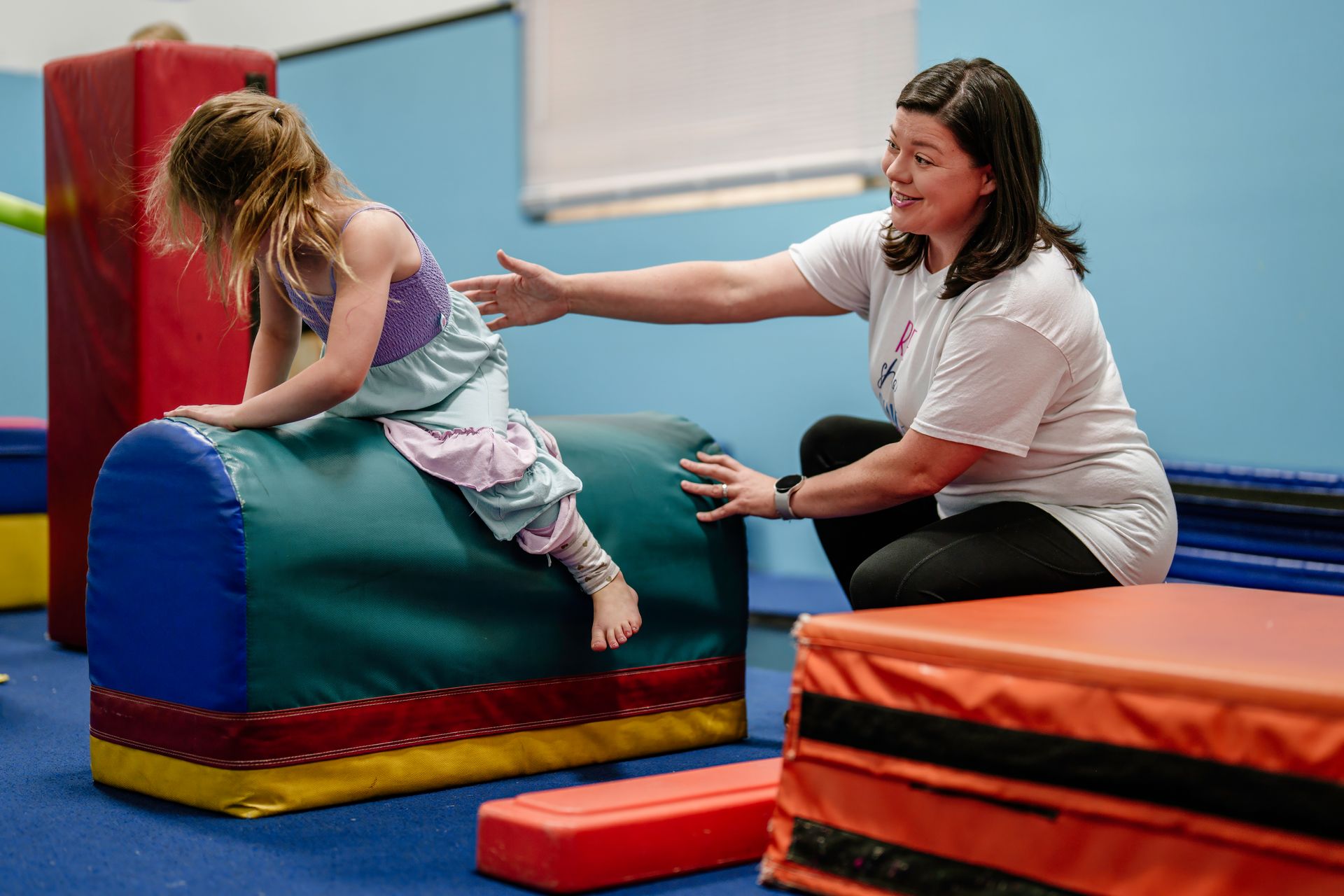 Child climbing blue foam block while smiling instructor helps in indoor gym with colorful mats