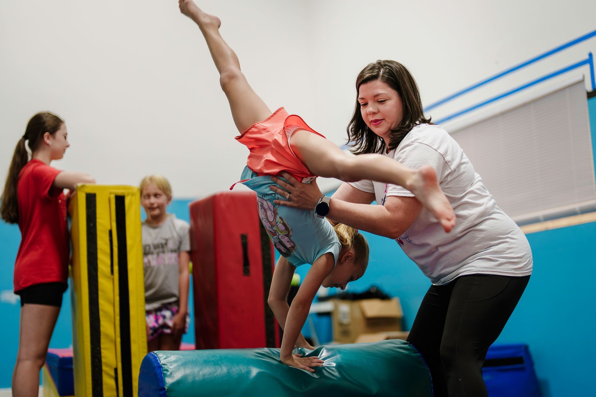 Child doing a handstand on a blue foam block while an instructor steadies them in a gymnasium