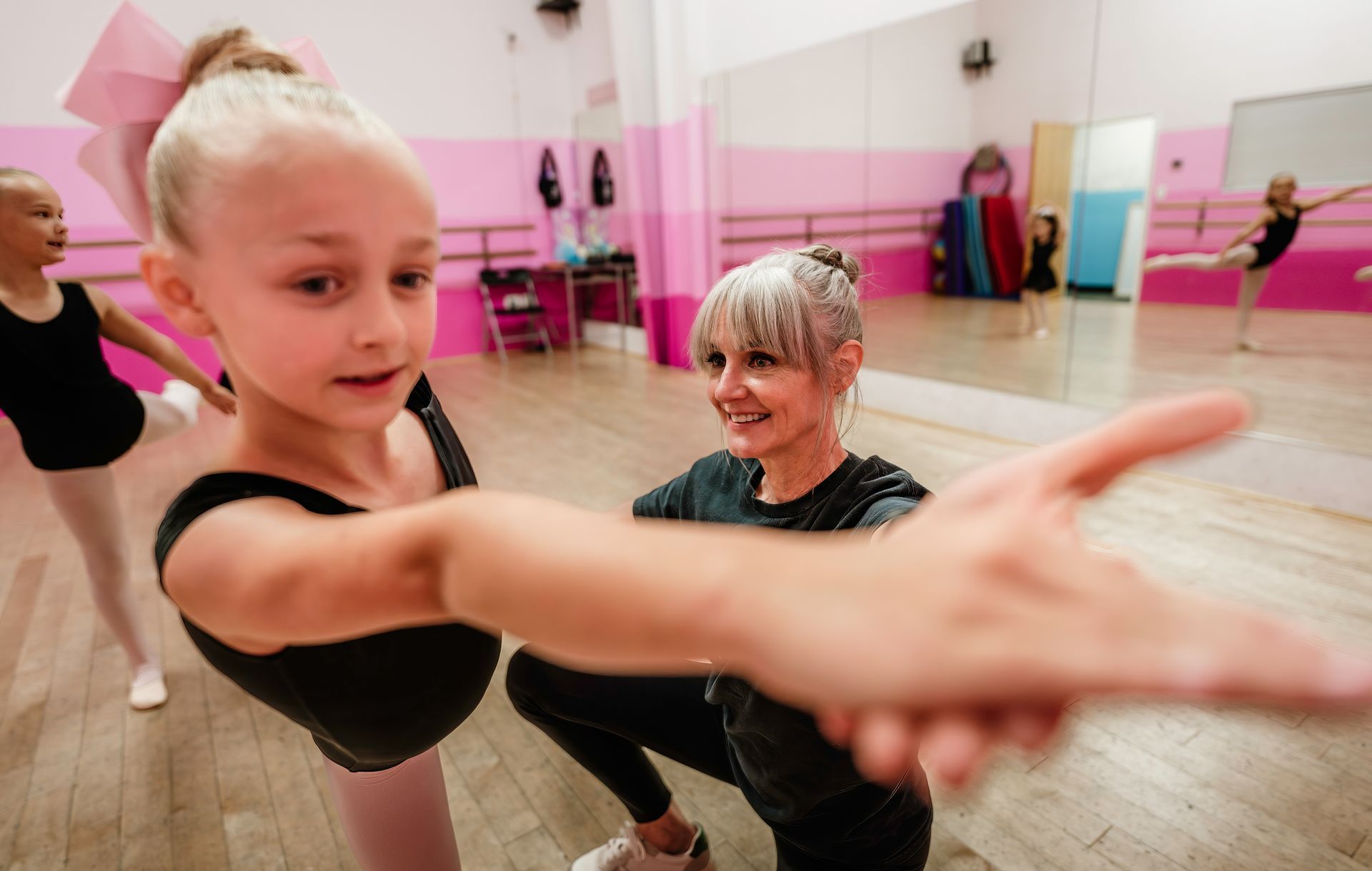 Dance class in a pink studio, with a girl stretching toward the camera and an instructor smiling behind her