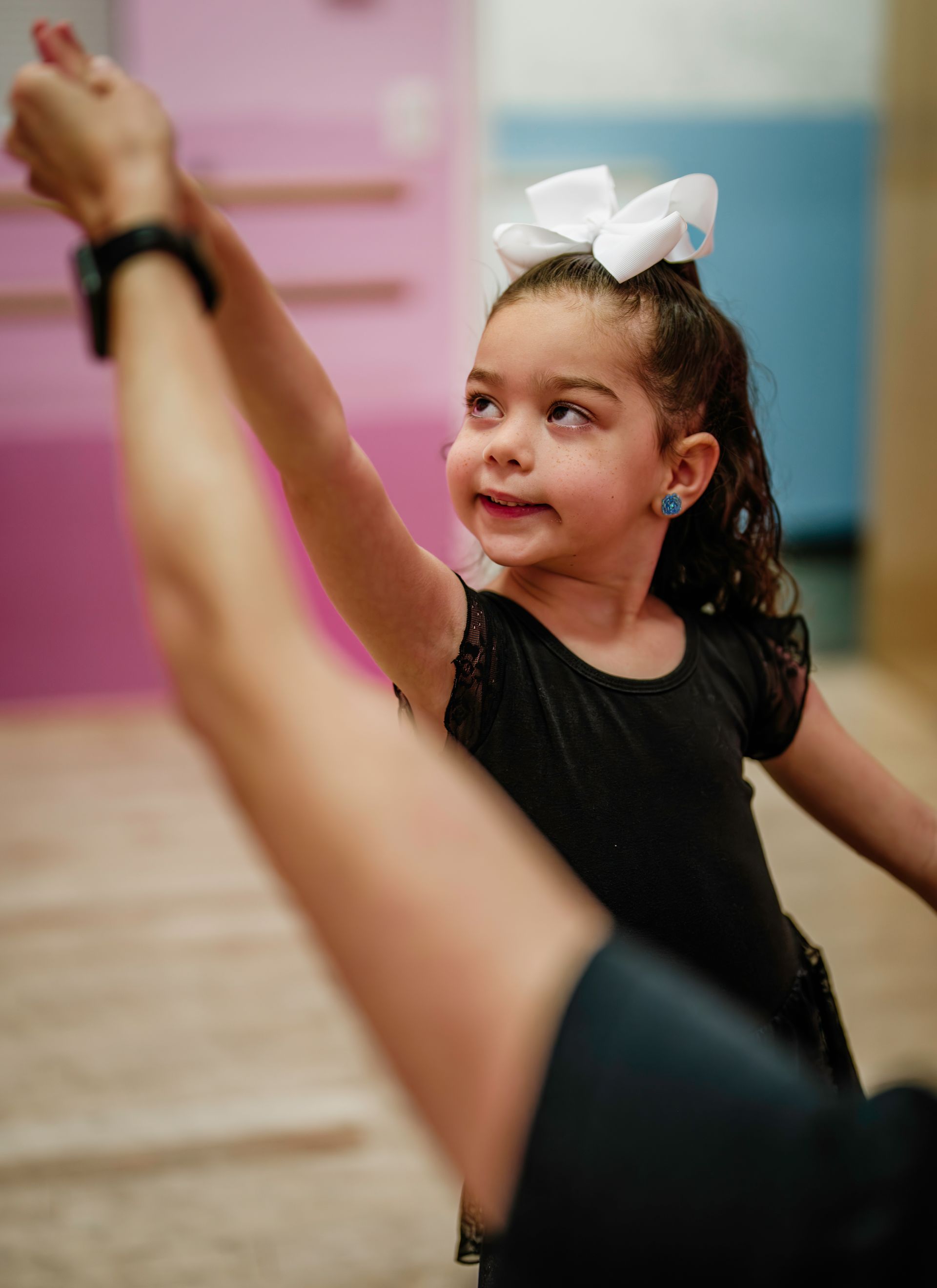 Young child in black dress with white bow, raising an arm in a dance studio, smiling at a teacher.